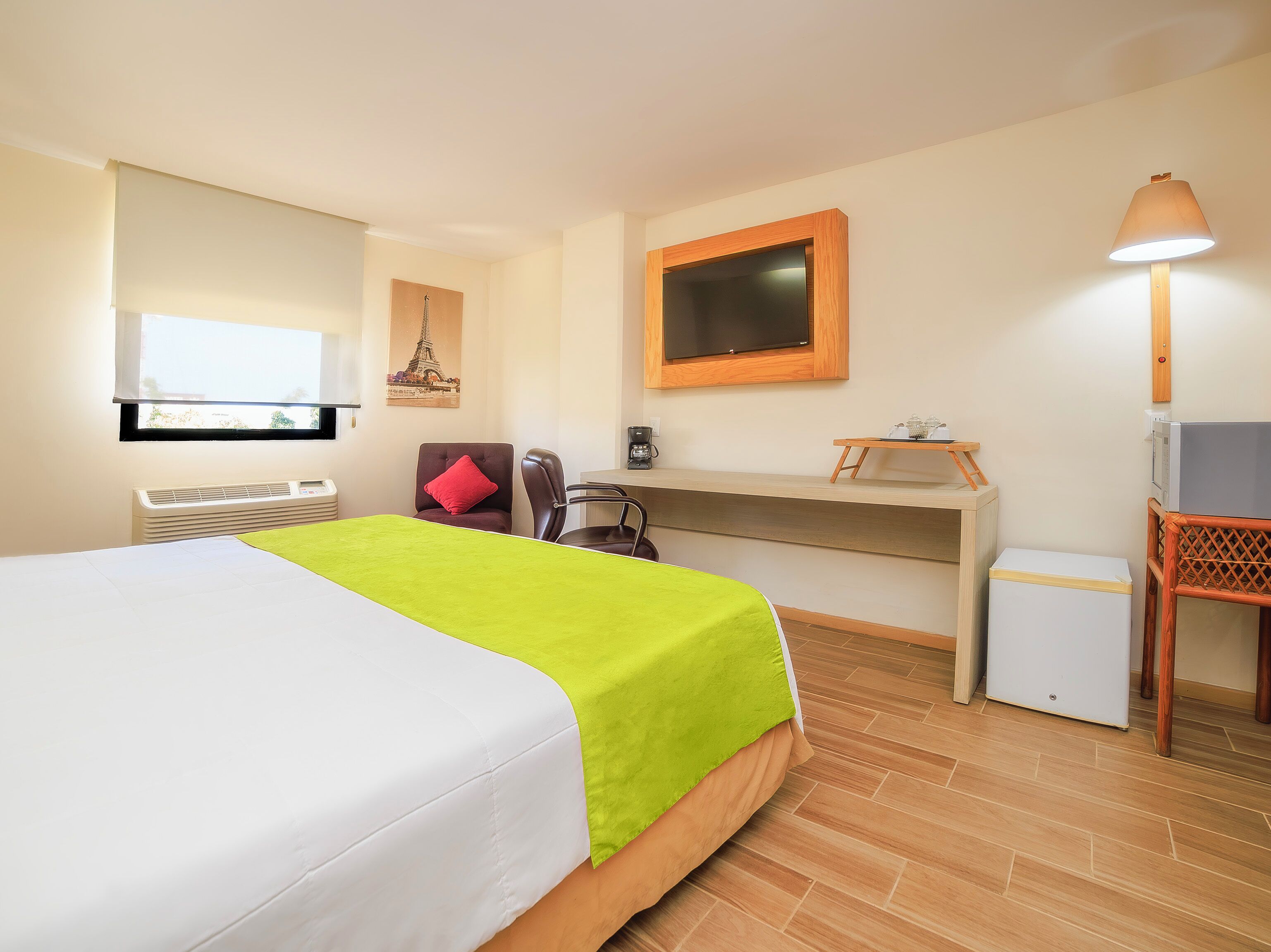 A hotel room with a bed, a desk, a chair, a small refrigerator, a heater, a window with a view, and a lamp.