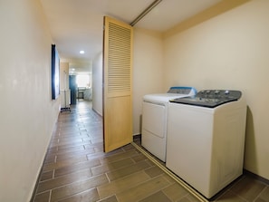 A laundry room with a washing machine and dryer, wooden door, tiled floor, and a hallway leading to a room with a bed and window.