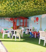 Uma área externa com mesas e cadeiras de plástico branco, uma estrutura com telhado vermelho e um mural com decorações temáticas de Paris.