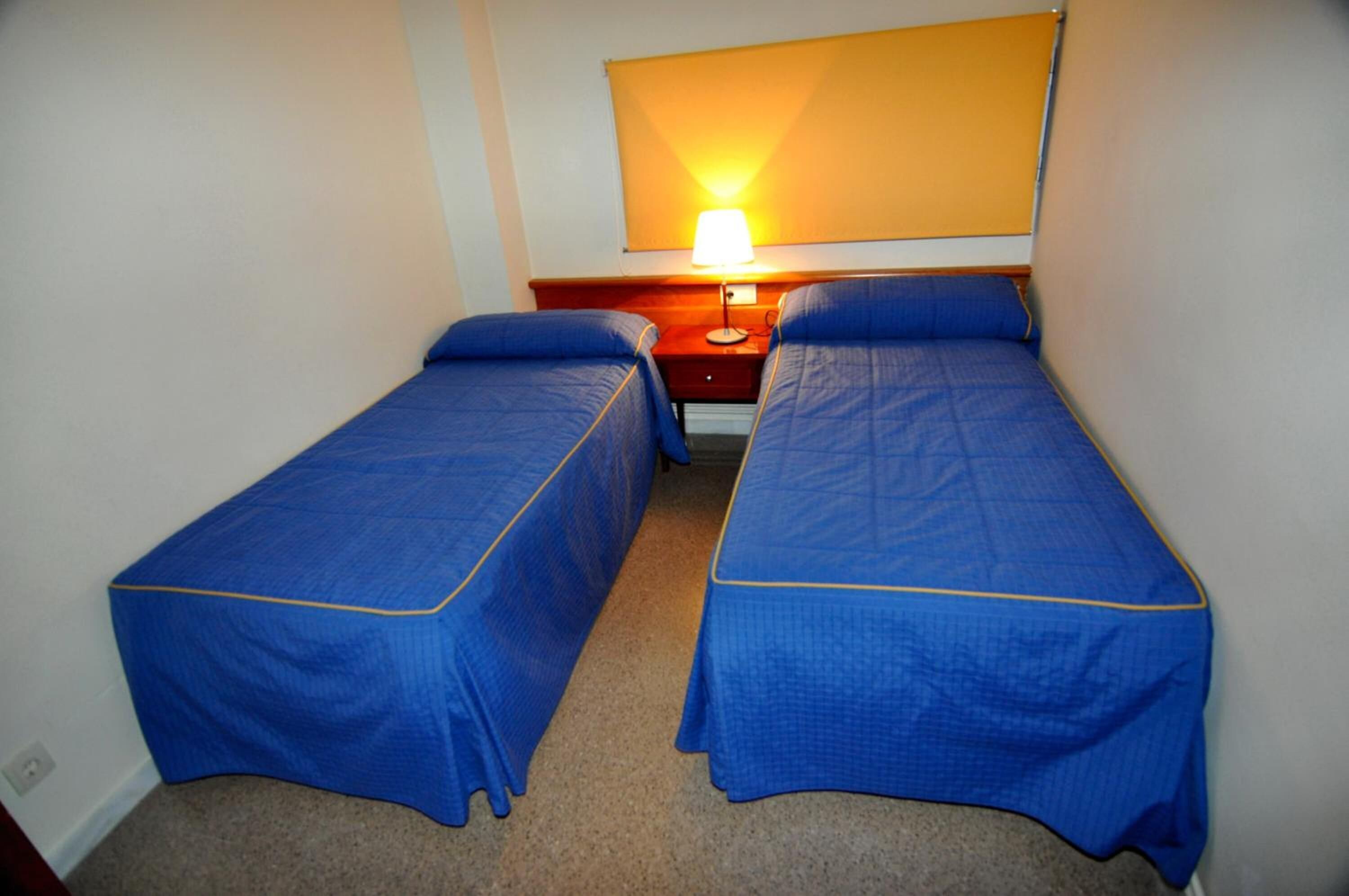 Una habitación pequeña y limpia con dos camas individuales, una mesita de noche con lámpara y un estante en la pared.