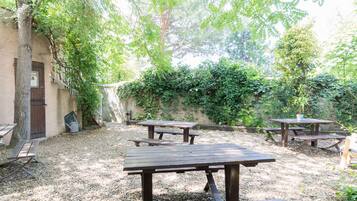 Un patio al aire libre con mesas y bancos de madera, rodeado de follaje y un muro de piedra.