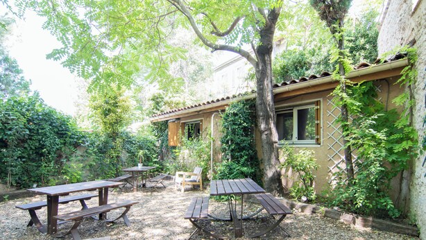 An outdoor patio area with picnic tables and benches, surrounded by trees and a building with a lattice window. - Résidence Carpe Diem (Marseille)