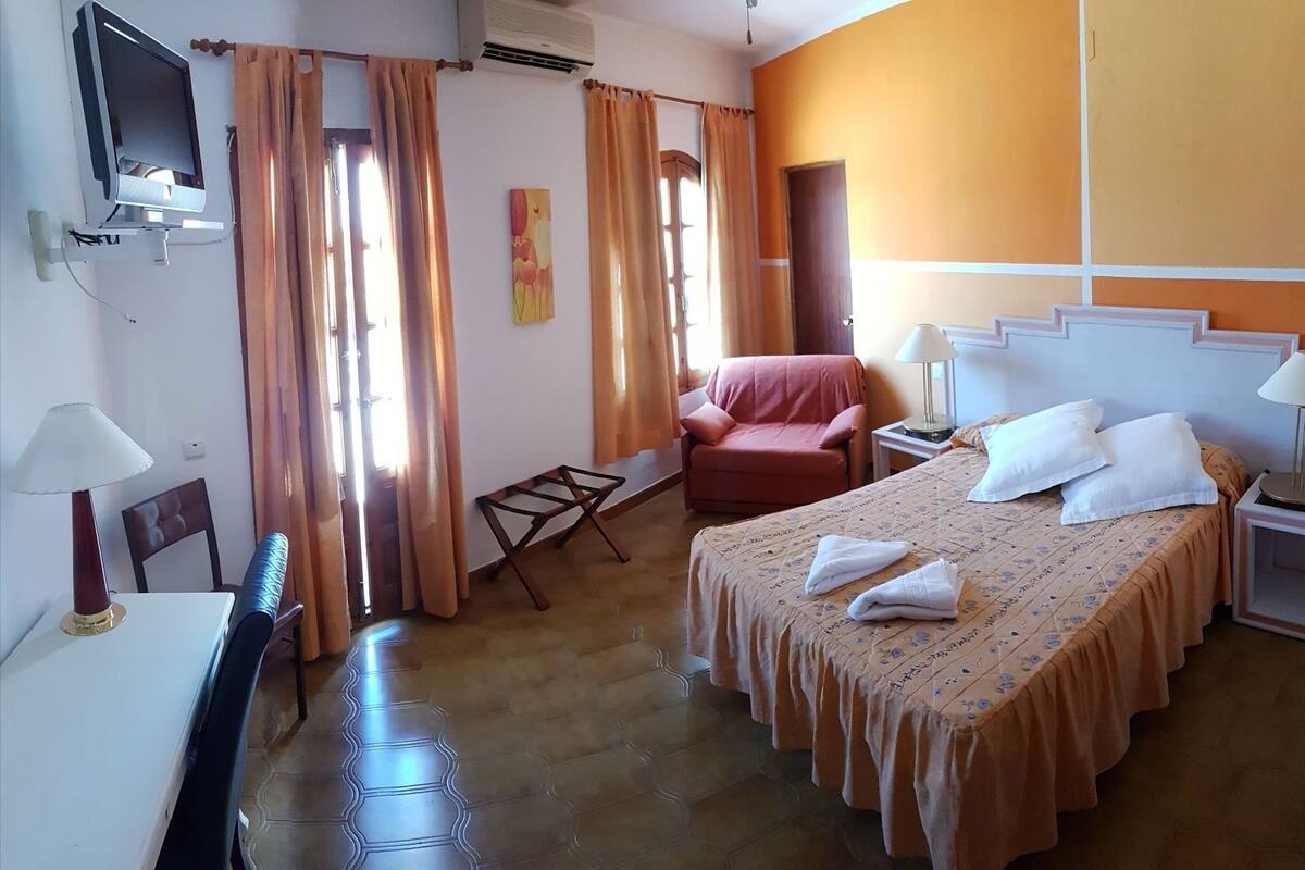 Quarto de hotel com cama, cadeira, escrivaninha, televisão e janela com cortinas.