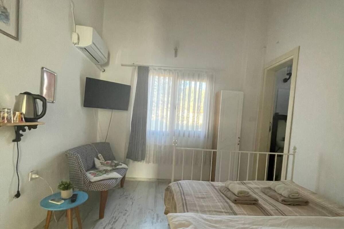 A small, well-lit room with a bed, a chair, a small table, and a TV mounted on the wall.