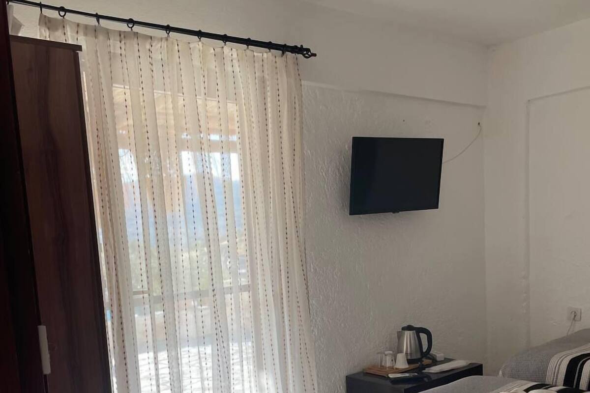 A bedroom with two beds, a TV mounted on the wall, a window with sheer curtains, a small bedside table with a kettle, and a red chair.