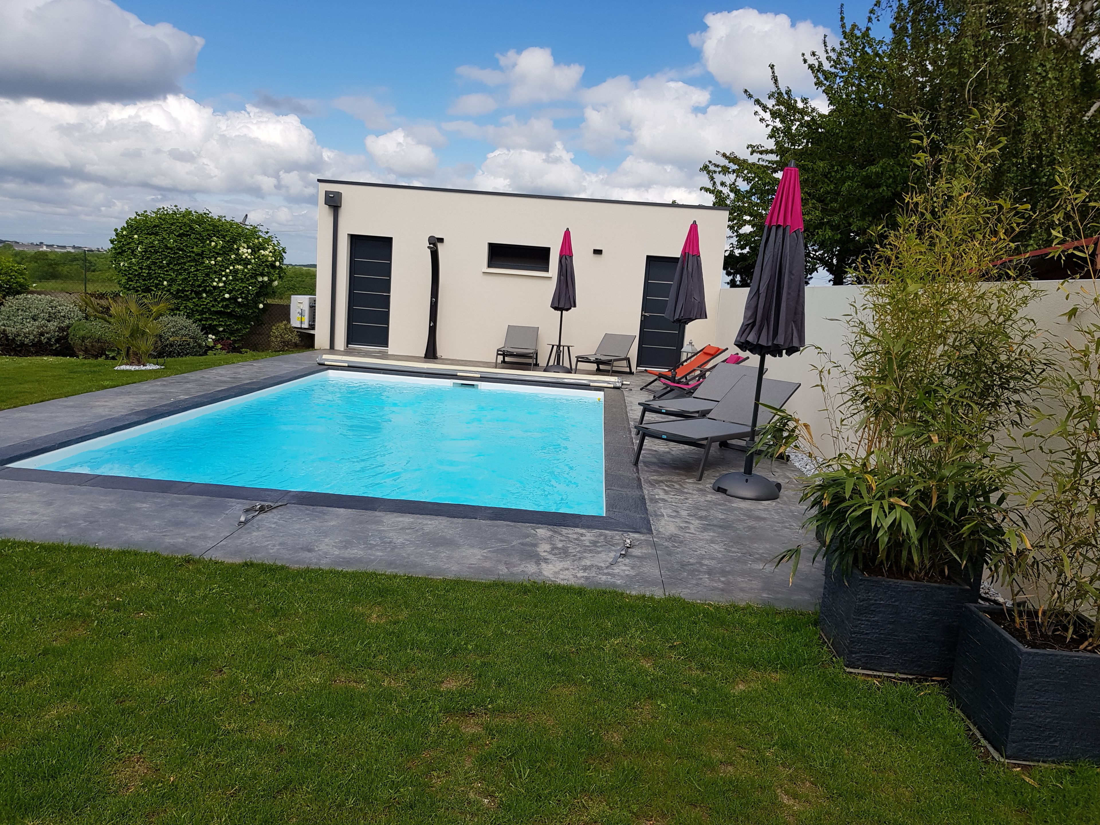 A rectangular swimming pool with a surrounding paved area, a modern building with a flat roof, and a few lounge chairs and umbrellas.