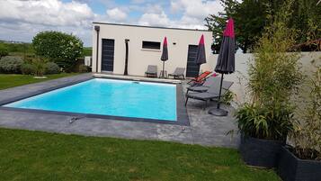 A rectangular swimming pool with a surrounding paved area, a modern building with a flat roof, and a few lounge chairs and umbrellas.