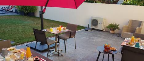 An outdoor patio area with tables and chairs, covered by a large red umbrella, and an air conditioning unit in the background.
