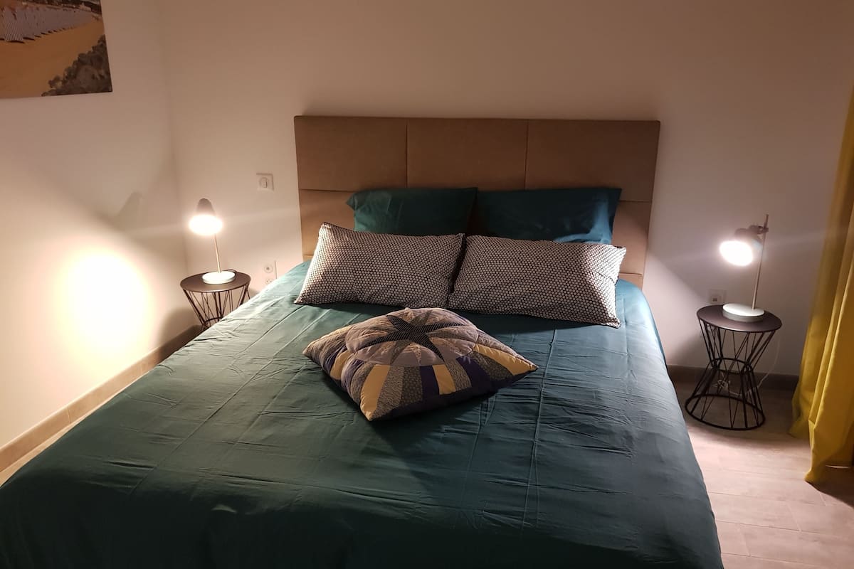A neatly made bed with a teal bedspread, two patterned pillows, and a quilted throw. There are two bedside lamps on either side of the bed.