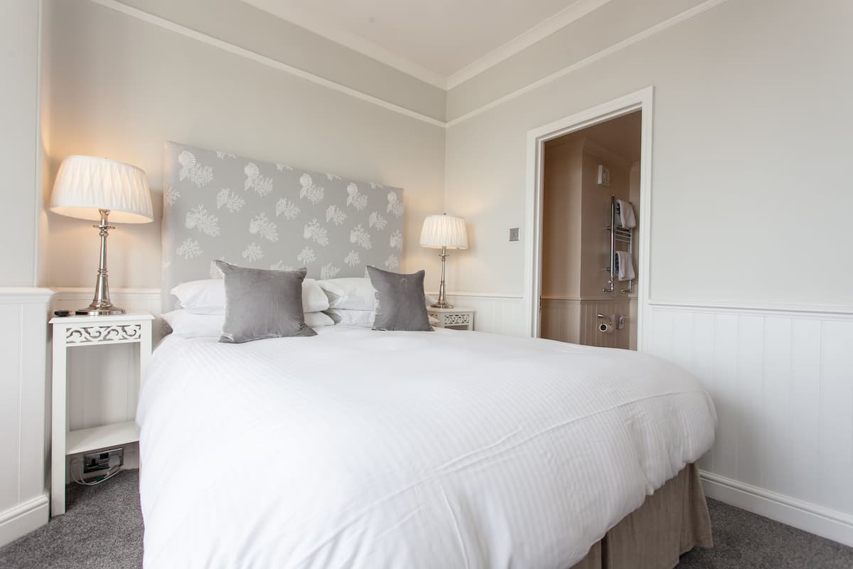 Een keurig opgemaakte slaapkamer met een groot bed, twee nachtkastjes met lampen, een kledingkast en een deur naar een andere kamer.