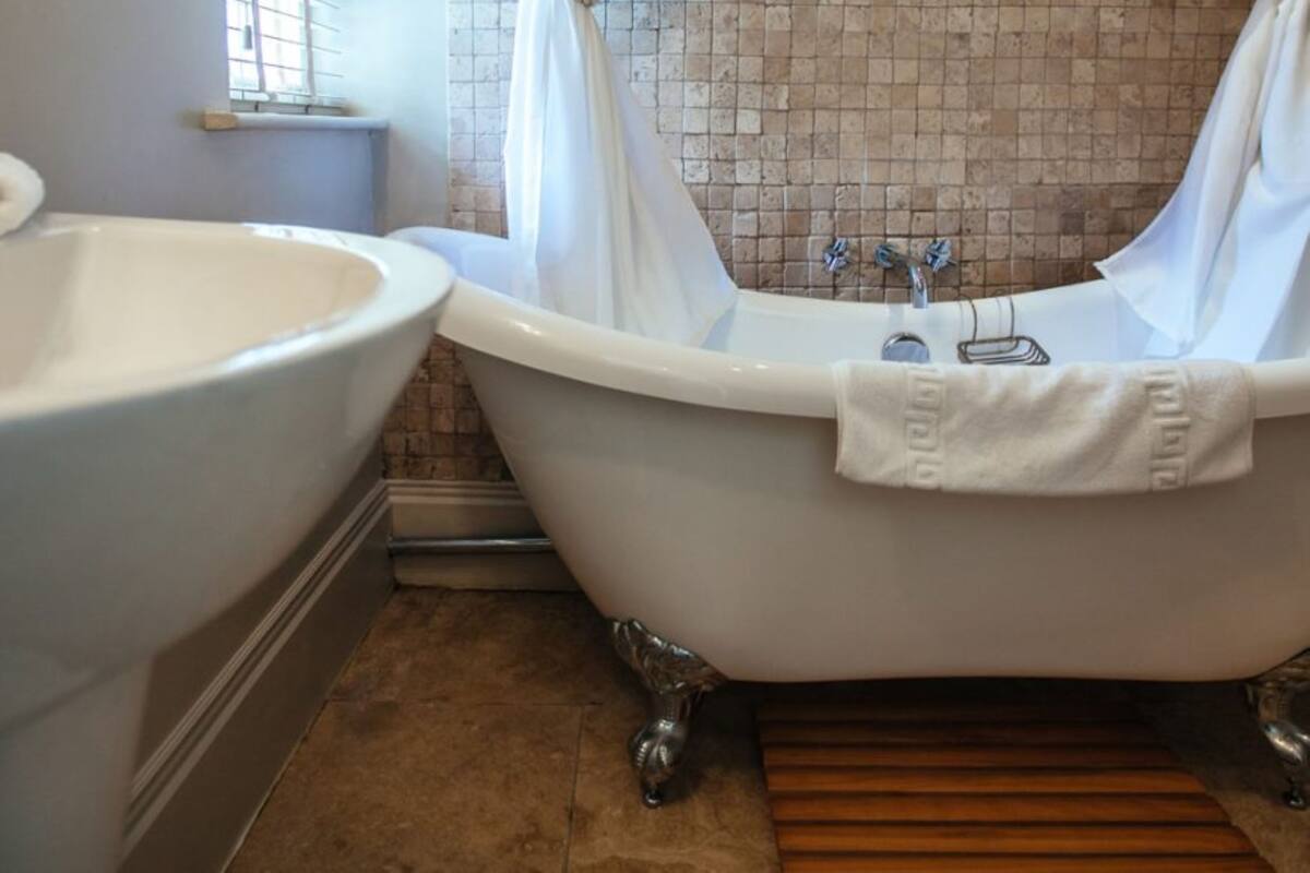 Een badkamer met een pootbad, een wastafel op een zuil en een raam met zonwering.