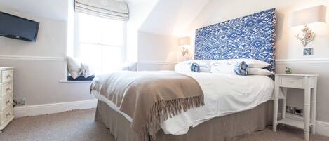 A bedroom with a bed, a nightstand, a lamp, a TV mounted on the wall, and a window with a view of the outside.
