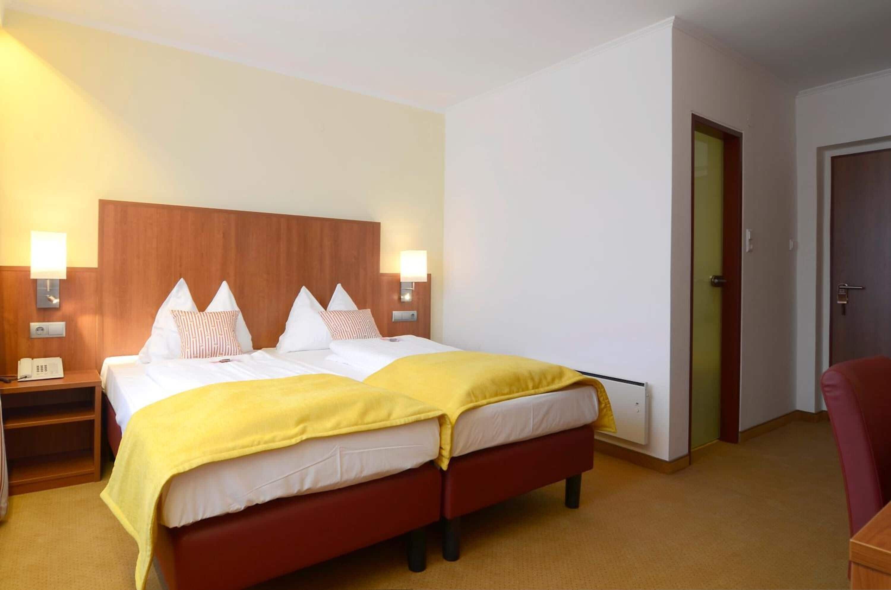 A hotel room with a double bed, two bedside tables with lamps, a wooden headboard, a red chair, and a door to the right.