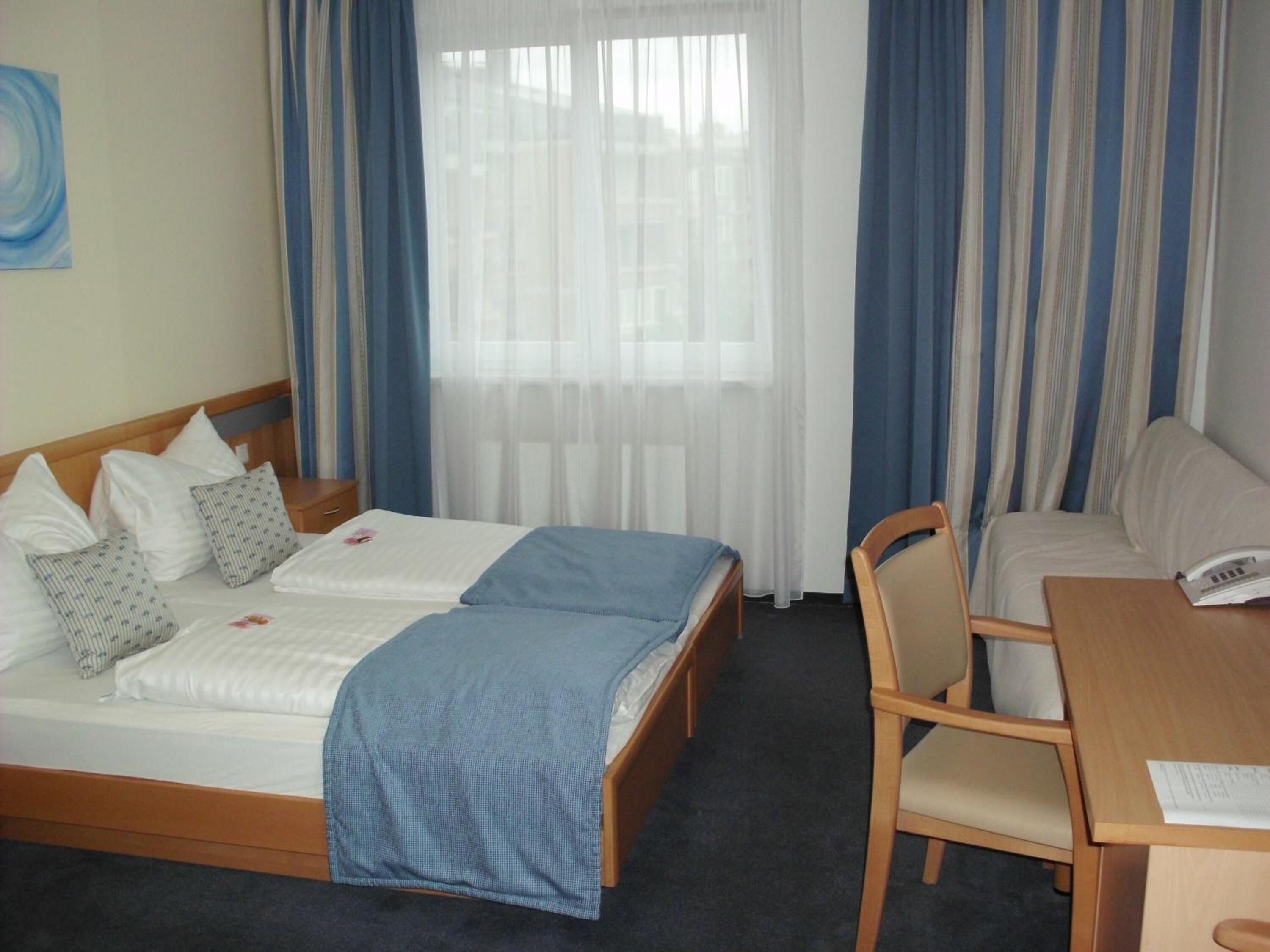A hotel room with two single beds, a wooden desk, a chair, and a window with sheer curtains.