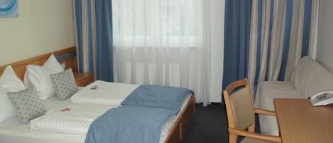A hotel room with two single beds, a wooden desk, a chair, and a window with sheer curtains.