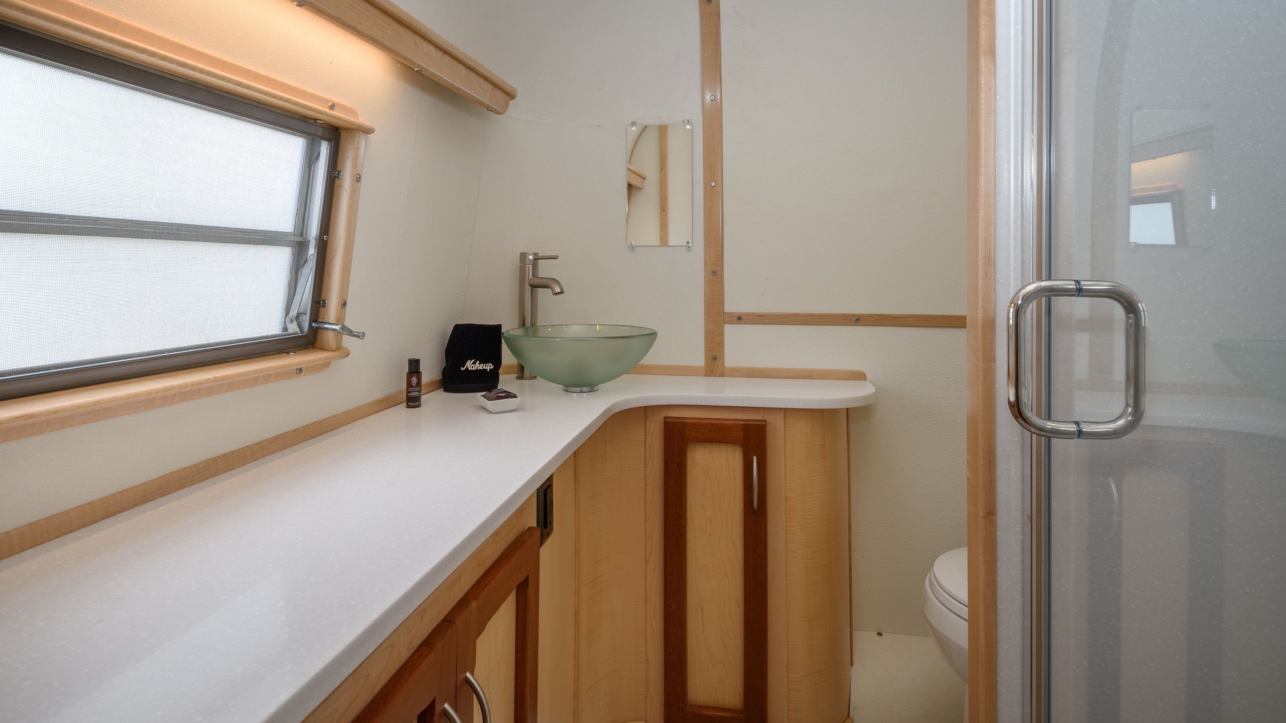 A compact bathroom with a window, a glass bowl sink, a makeup bag, and a toilet.