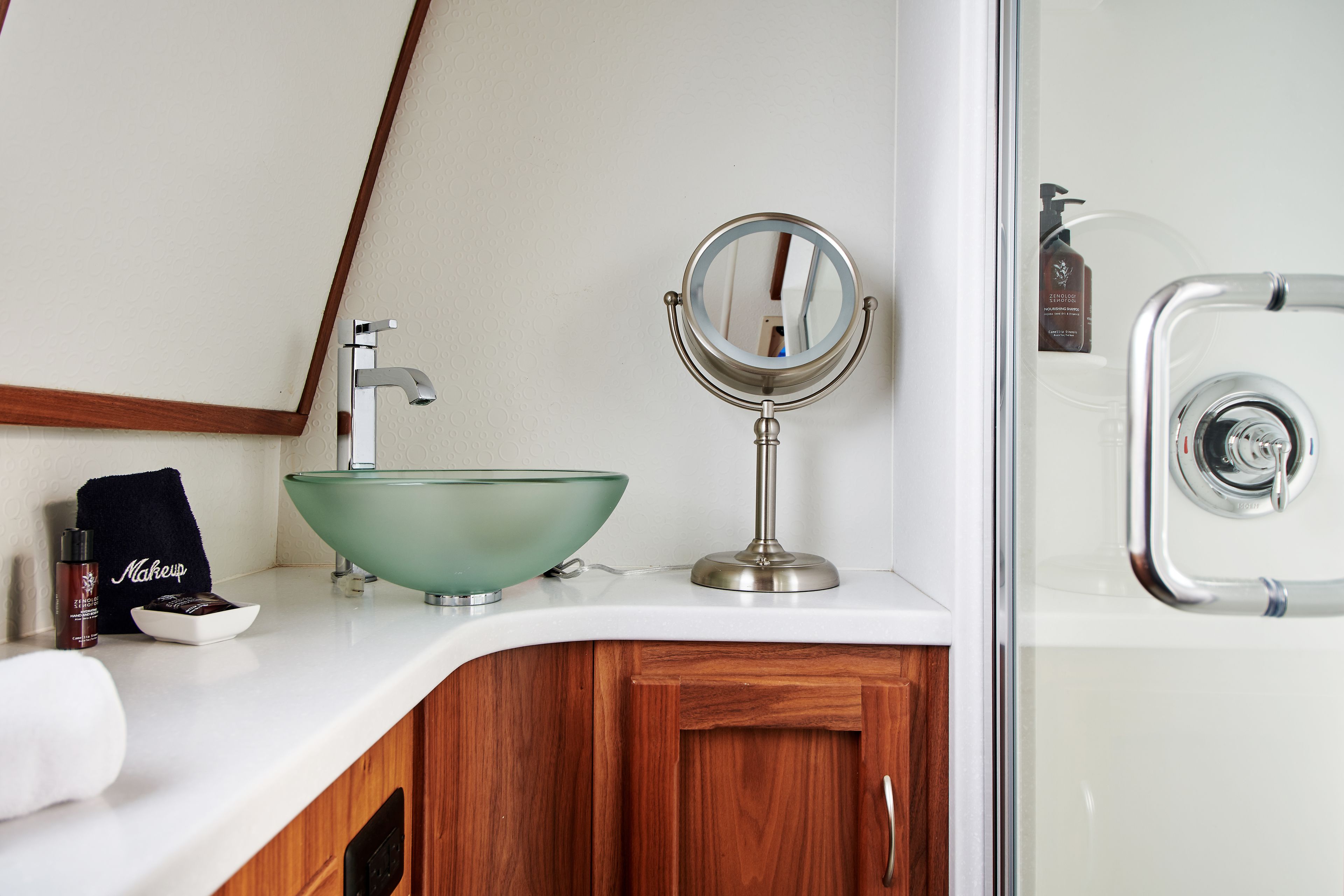 A bathroom with a white countertop, a green glass bowl sink, a round mirror, a makeup bag, and a bottle of shampoo.