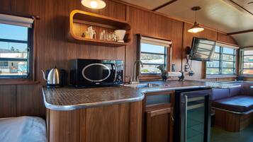 A compact kitchen with wooden cabinetry, a microwave, a kettle, and a sink. There are shelves above the counter and a television mounted on the wall.