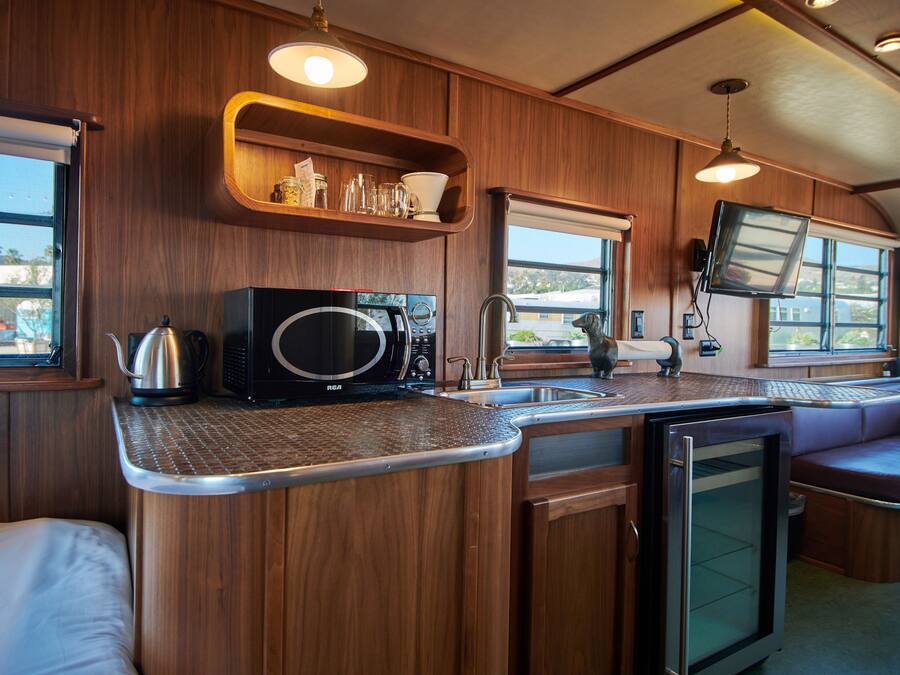 A compact kitchen with wooden cabinetry, a microwave, a kettle, and a sink. There are shelves above the counter and a television mounted on the wall.