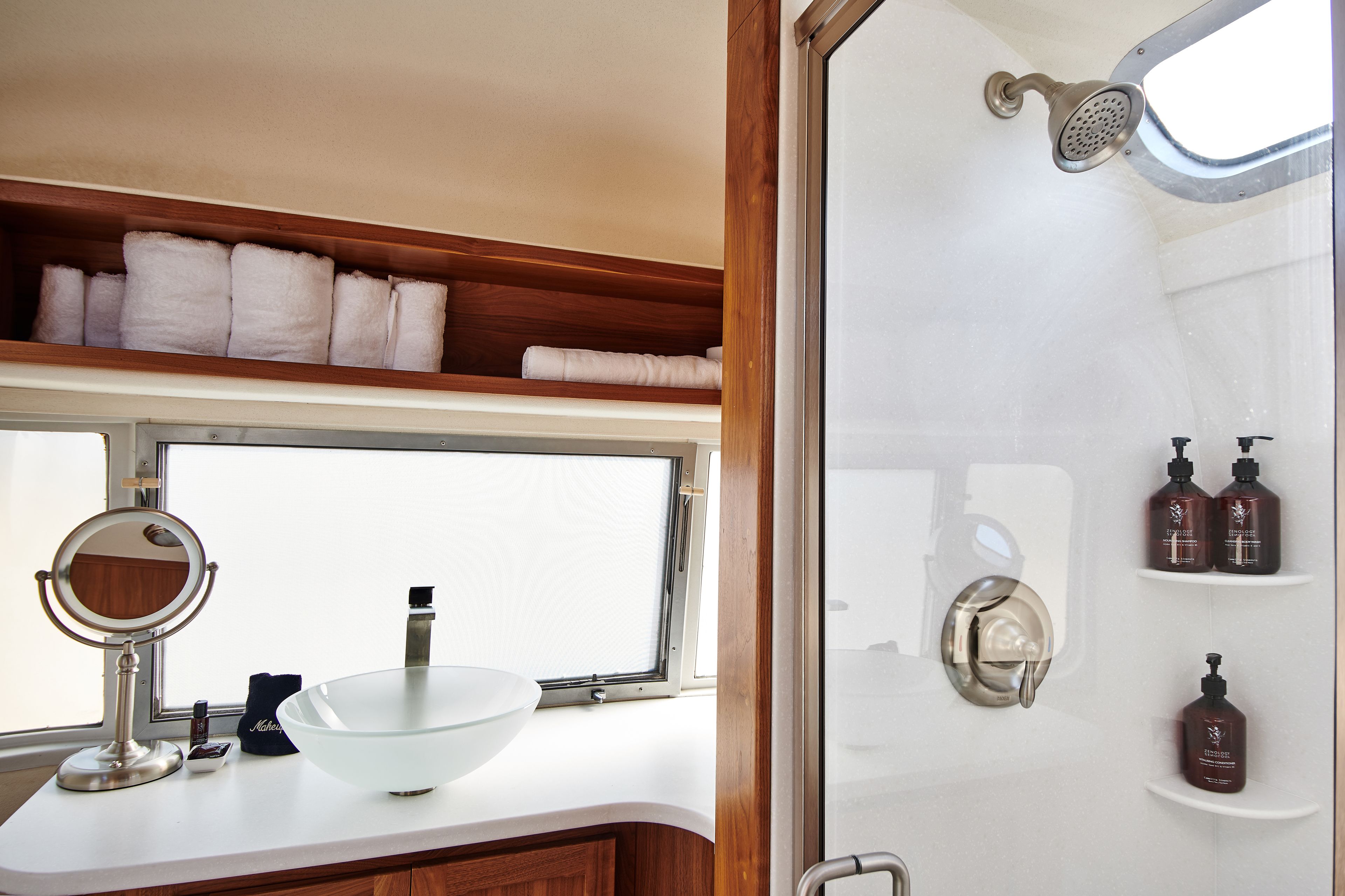 A bathroom with a large window, a sink with a mirror, a shower, and shelves with toiletries.