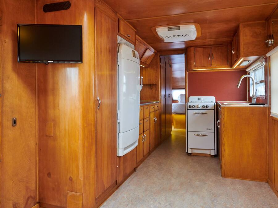 A compact kitchen with wooden cabinets, a white refrigerator, a stove, and a flat-screen TV mounted on the wall.