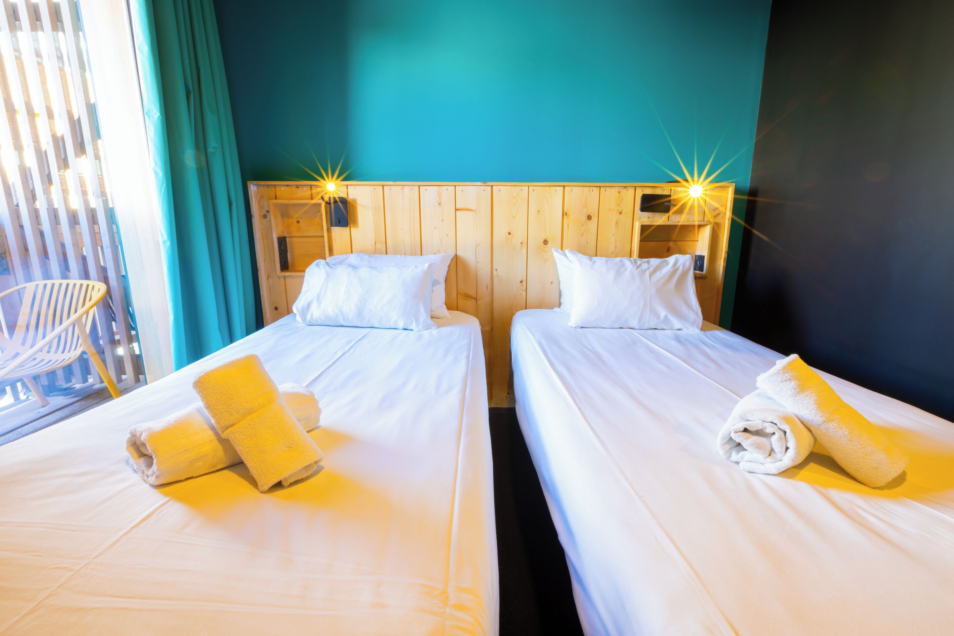 Een hotelkamer met twee bedden, elk voorzien van witte lakens en een opgerolde handdoek. Er is een houten hoofdbord met een lamp boven de bedden.