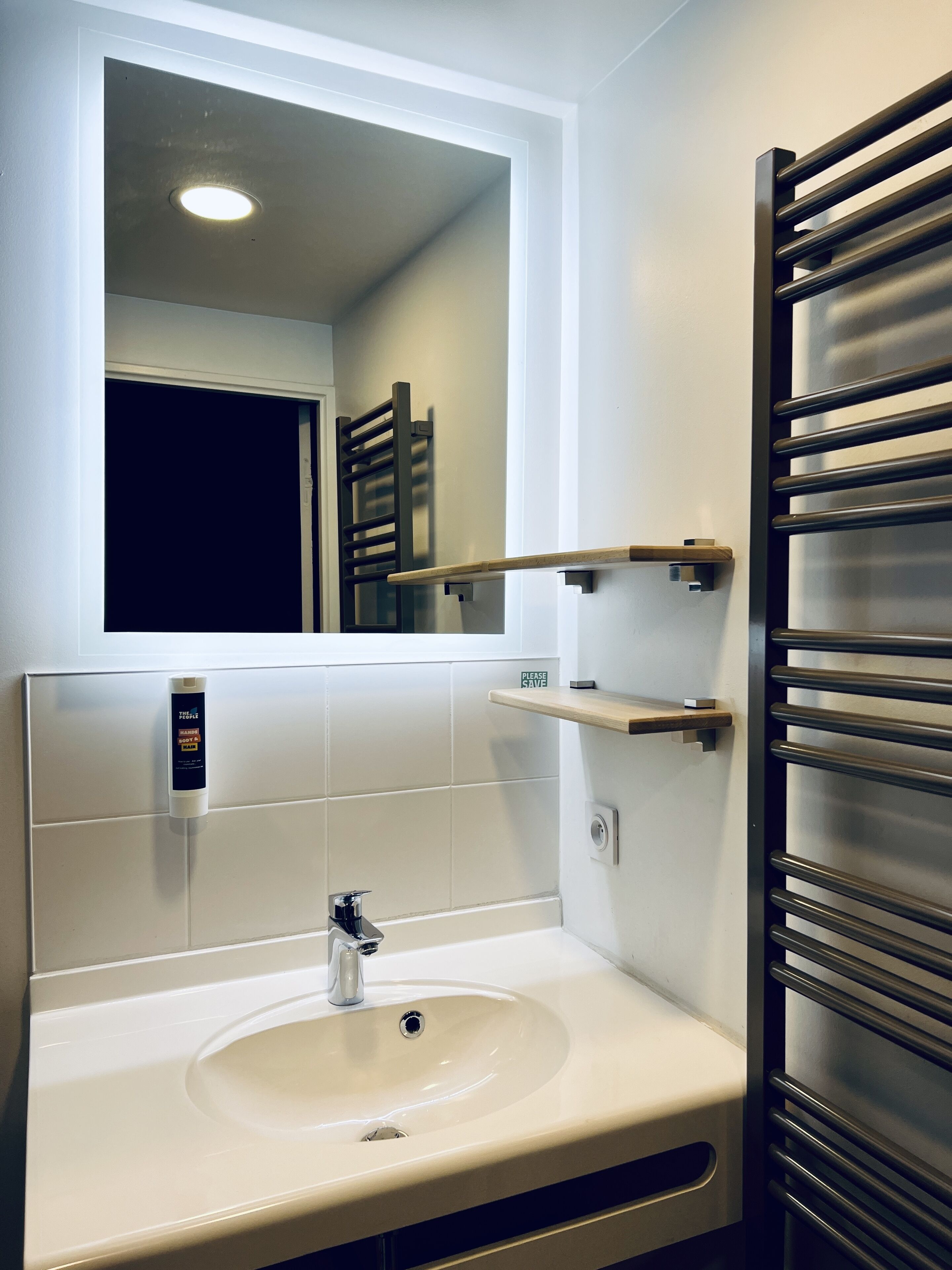Un bagno con un lavandino bianco, uno specchio, un porta asciugamani e uno scaffale.