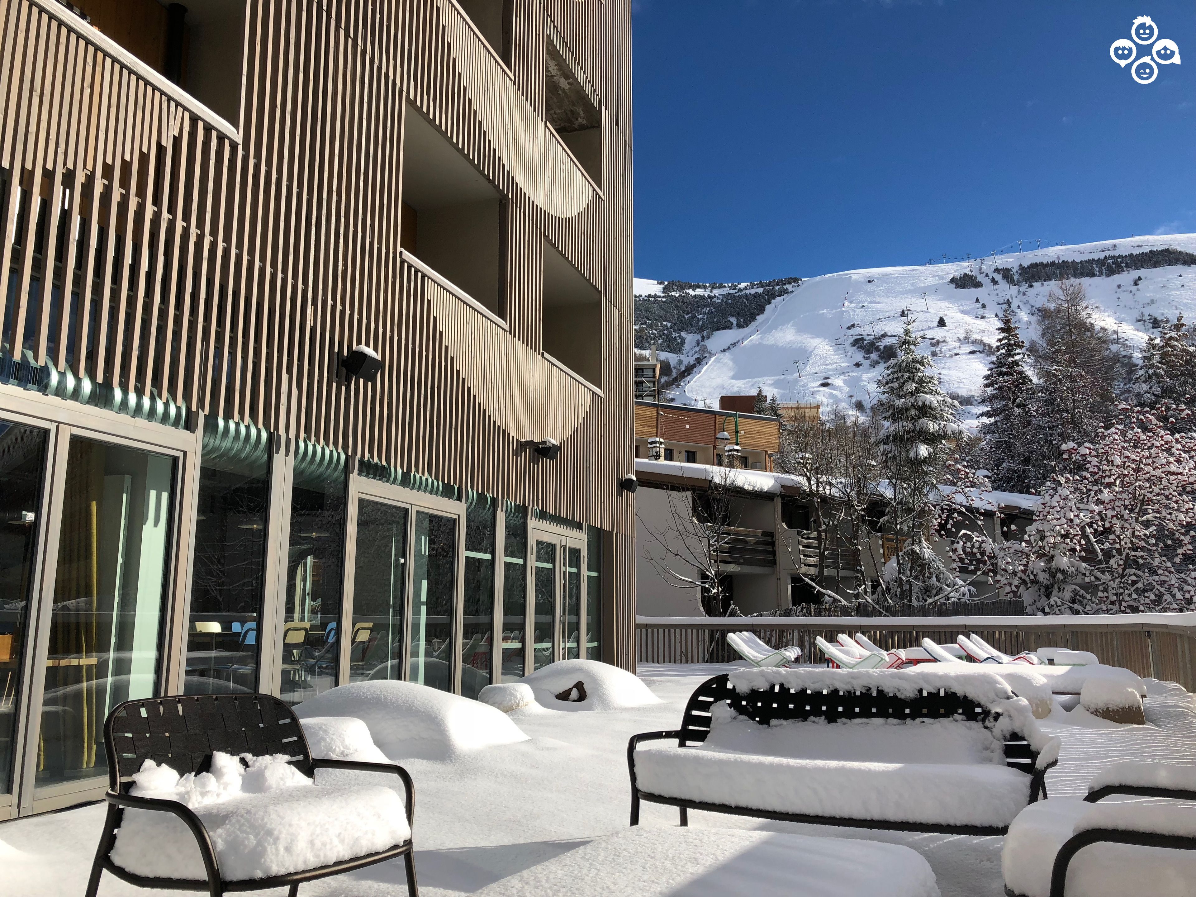 A snow-covered outdoor seating area with a view of a snow-covered mountain and a building with vertical wooden slats.