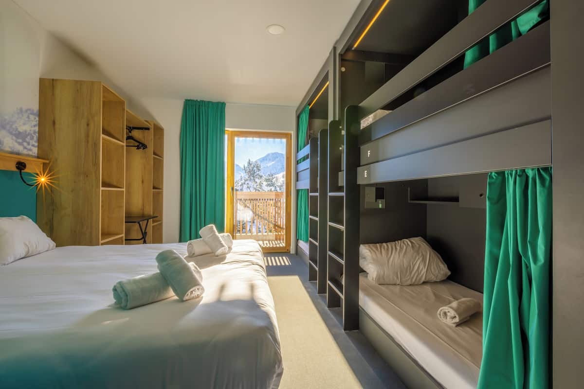 Une chambre d’hôtel avec un lit superposé, une armoire en bois et une grande fenêtre avec des rideaux.