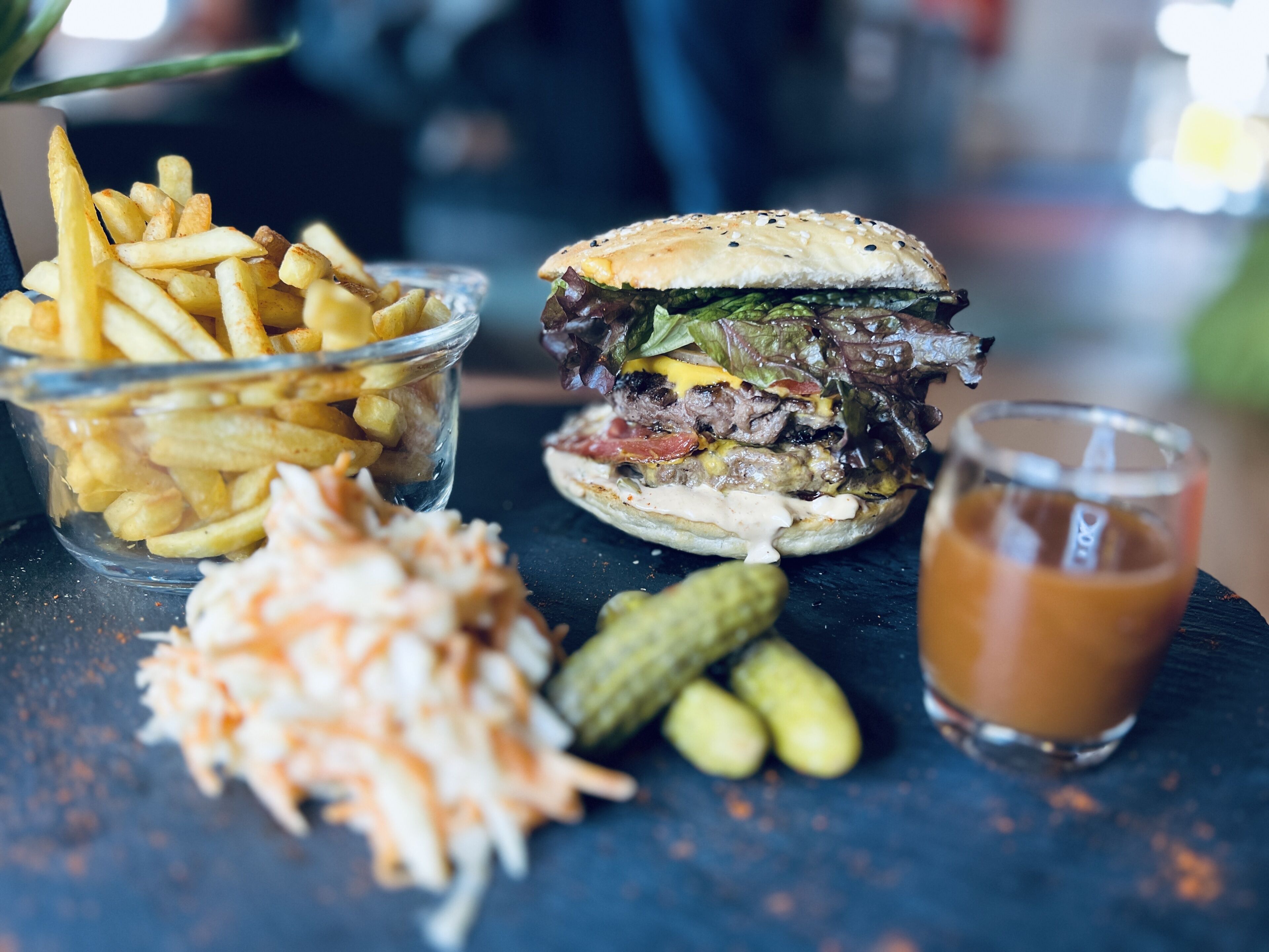 Een maaltijd bestaande uit een hamburger, frieten, een salade van kool, augurken en een drankje op een donker oppervlak.