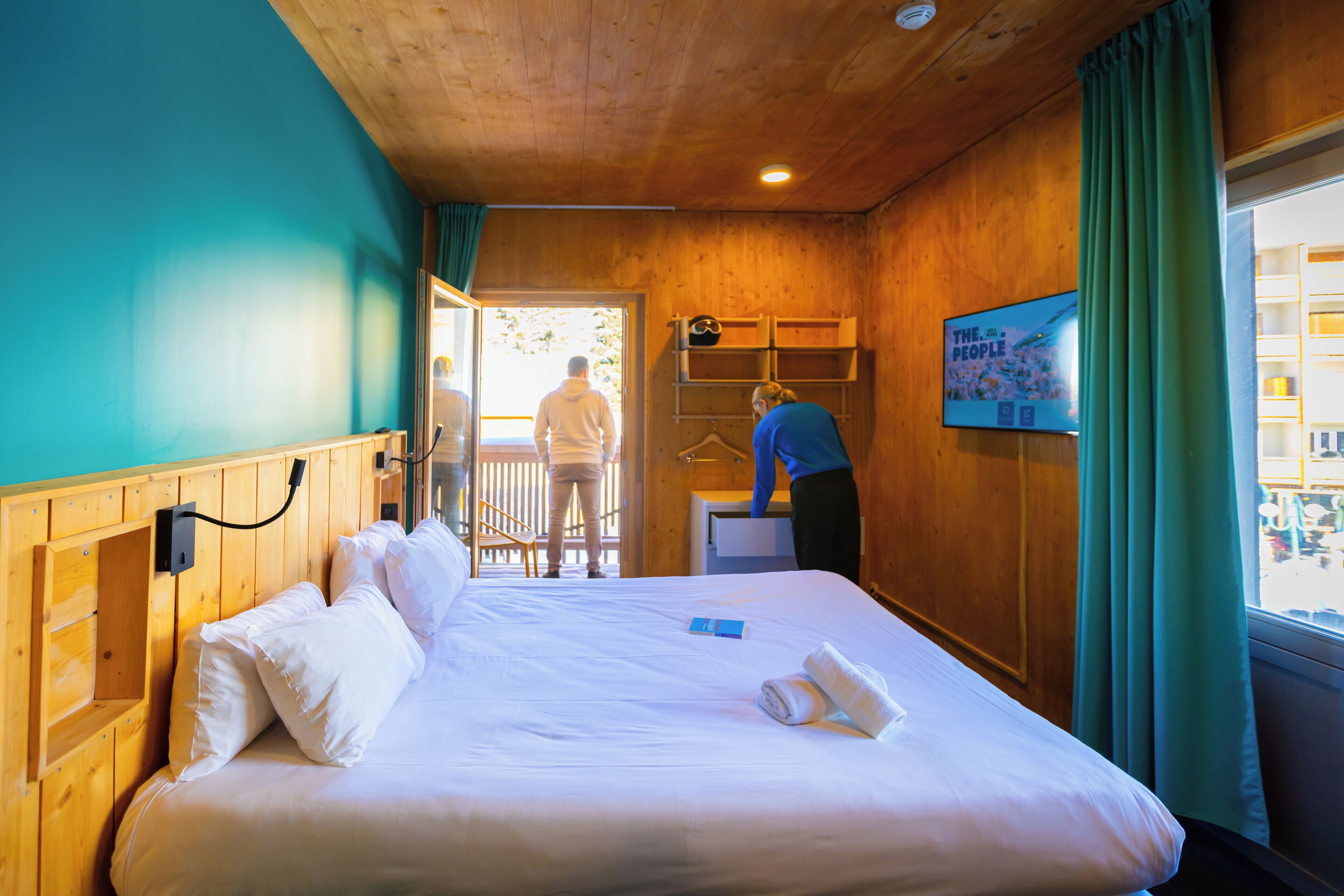 A hotel room with a wooden ceiling, a bed with white linens, a small desk, and a window with curtains.
