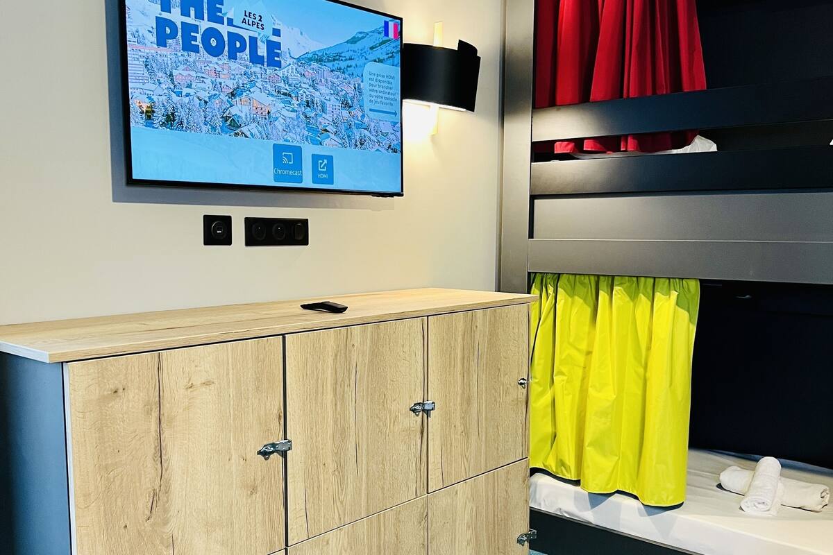 Une chambre avec un téléviseur diffusant l'émission « The People Les 2 Alpes », un meuble en bois, des lits superposés avec des rideaux rouges et jaunes, et une chaise jaune.