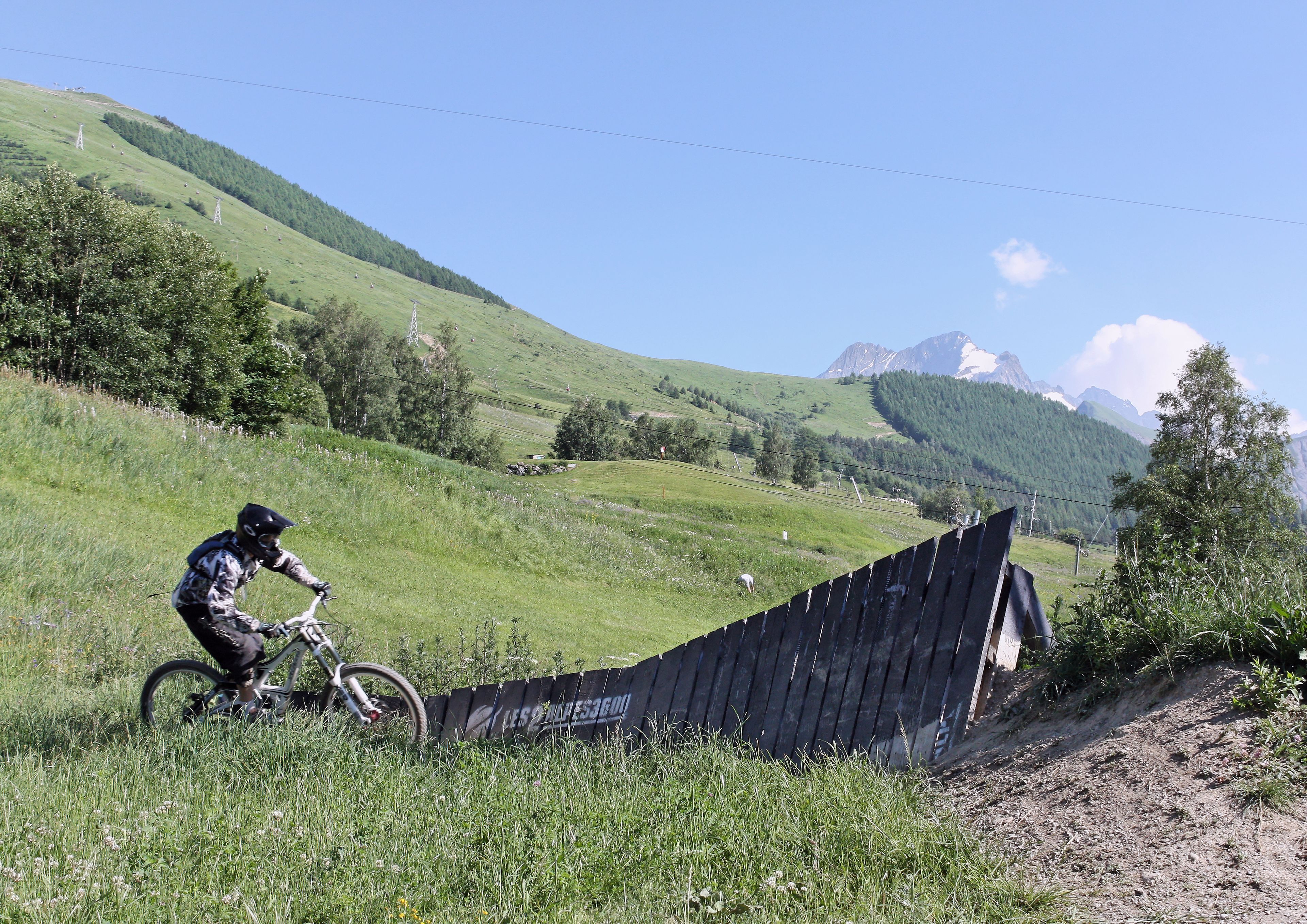 A person riding a mountain bike on a grassy slope with a wooden ramp.