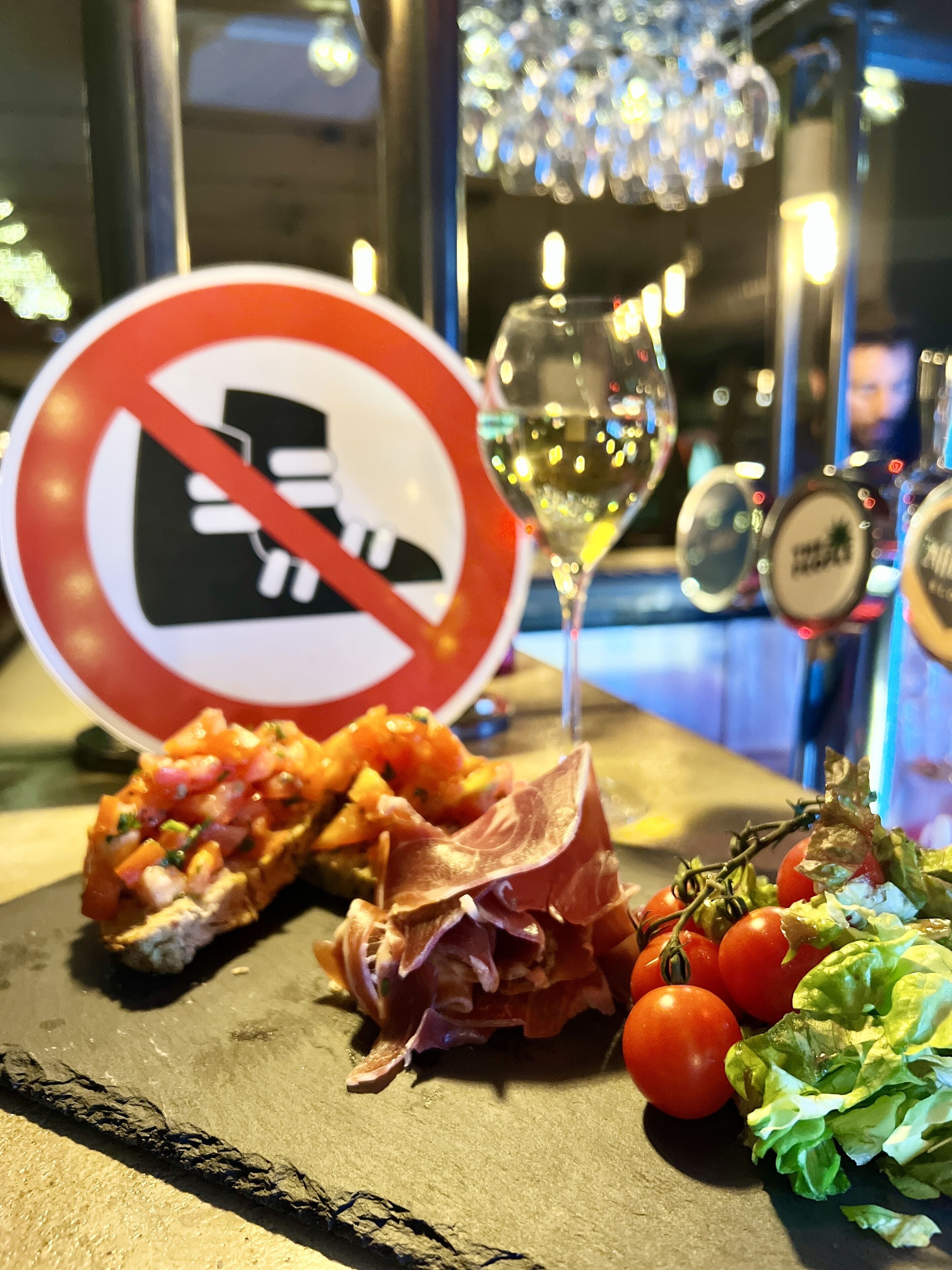 Un bar avec un panneau indiquant qu’il est interdit de fumer, un verre de vin blanc et une assiette de nourriture comprenant des bruschettas, du jambon de Parme et des tomates.