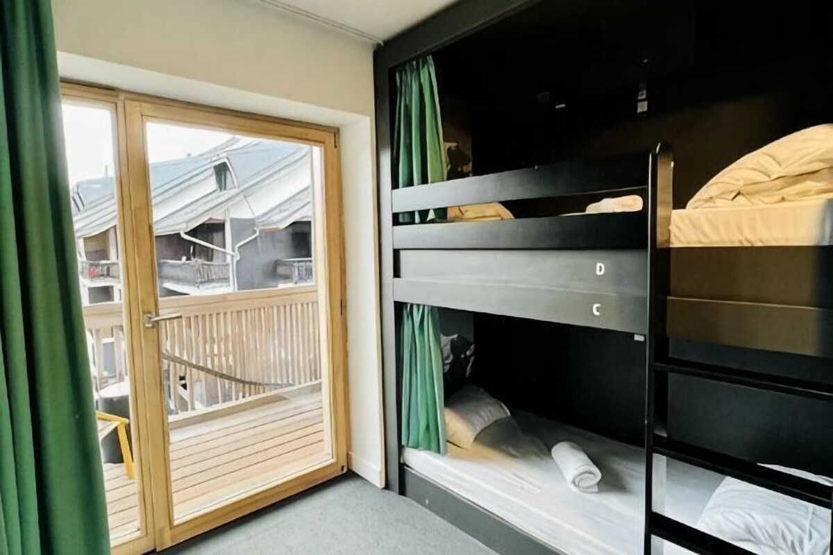 A room with bunk beds, a sliding glass door to a balcony, and a view of buildings outside.