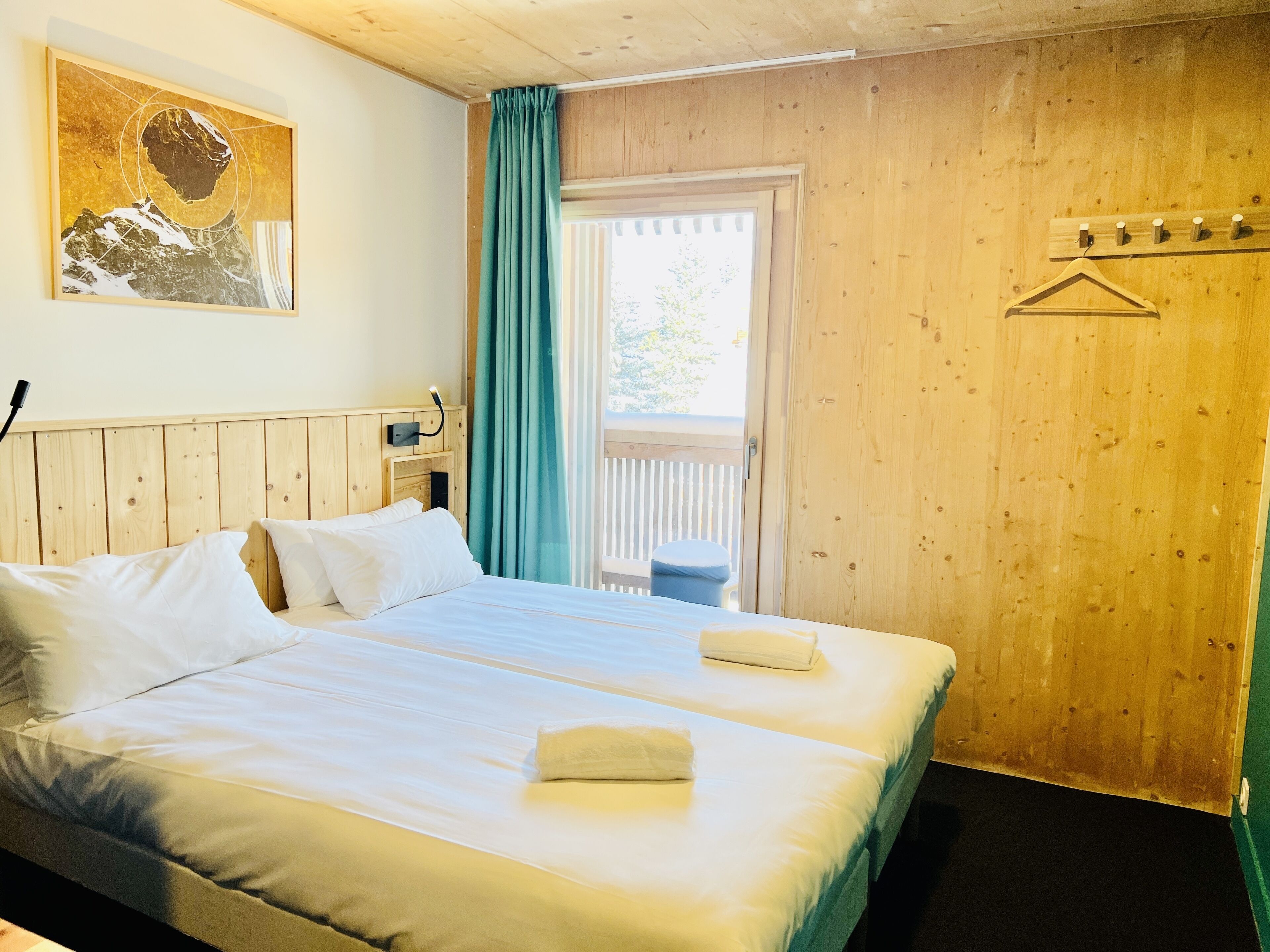 Ein Zimmer mit zwei Betten, einem Holzkopfende, einem Gemälde, einem Fenster mit Vorhängen und einem Balkon.