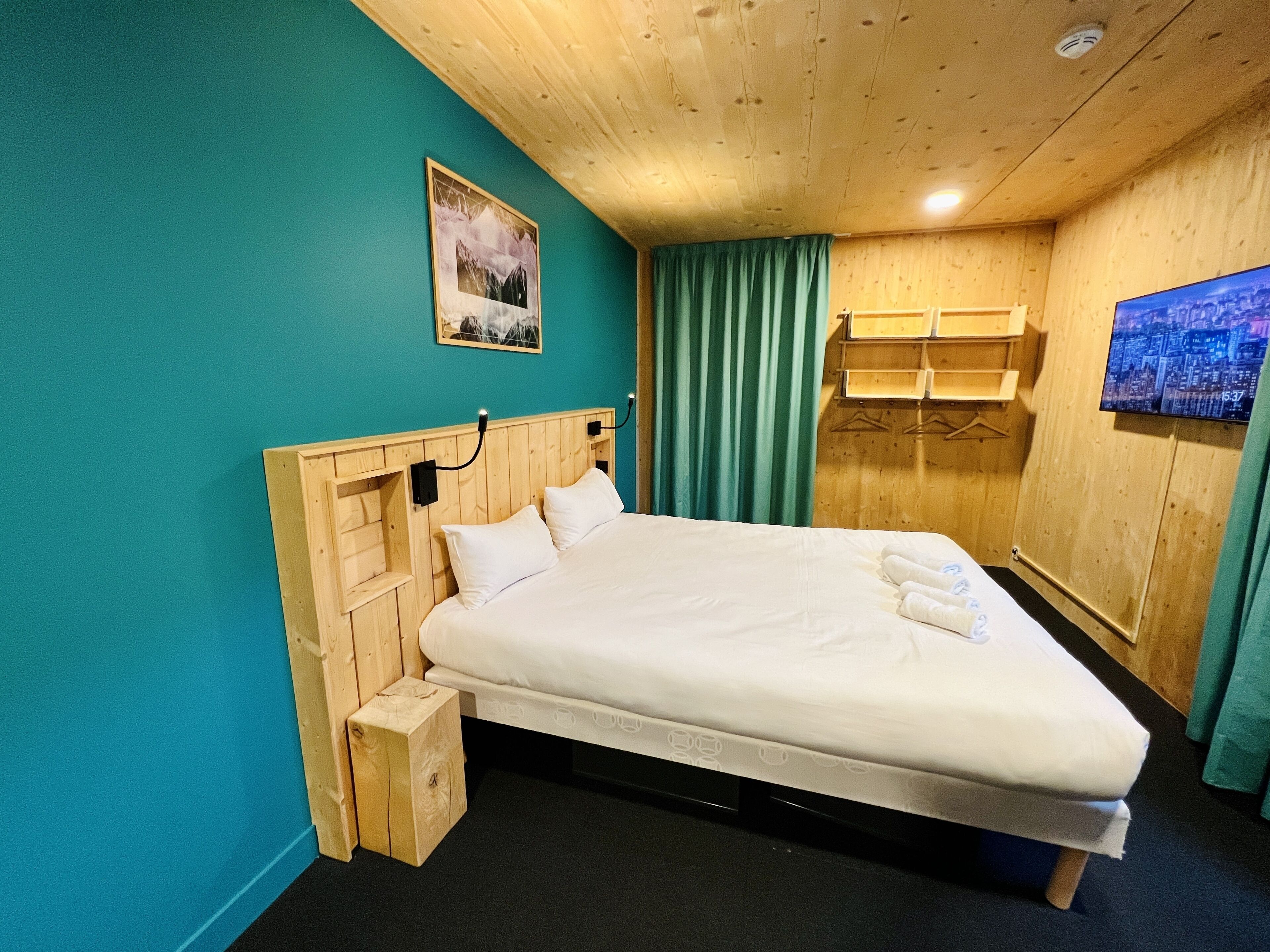 A wooden bed with a headboard, a small bedside table, a wall-mounted TV, and a window with curtains.