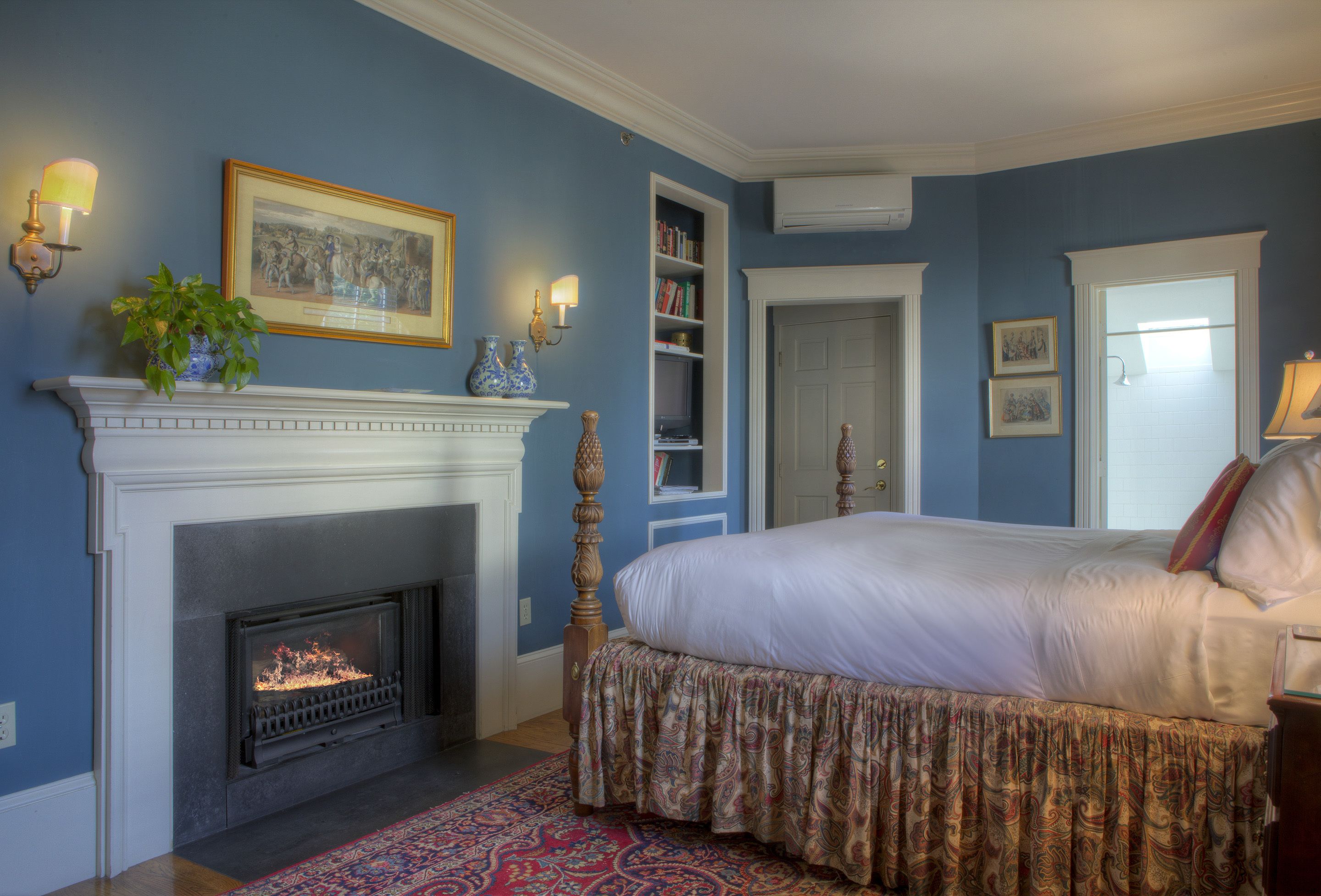 A bedroom with a fireplace, a bed with a patterned bedspread, a bookshelf, and a door.