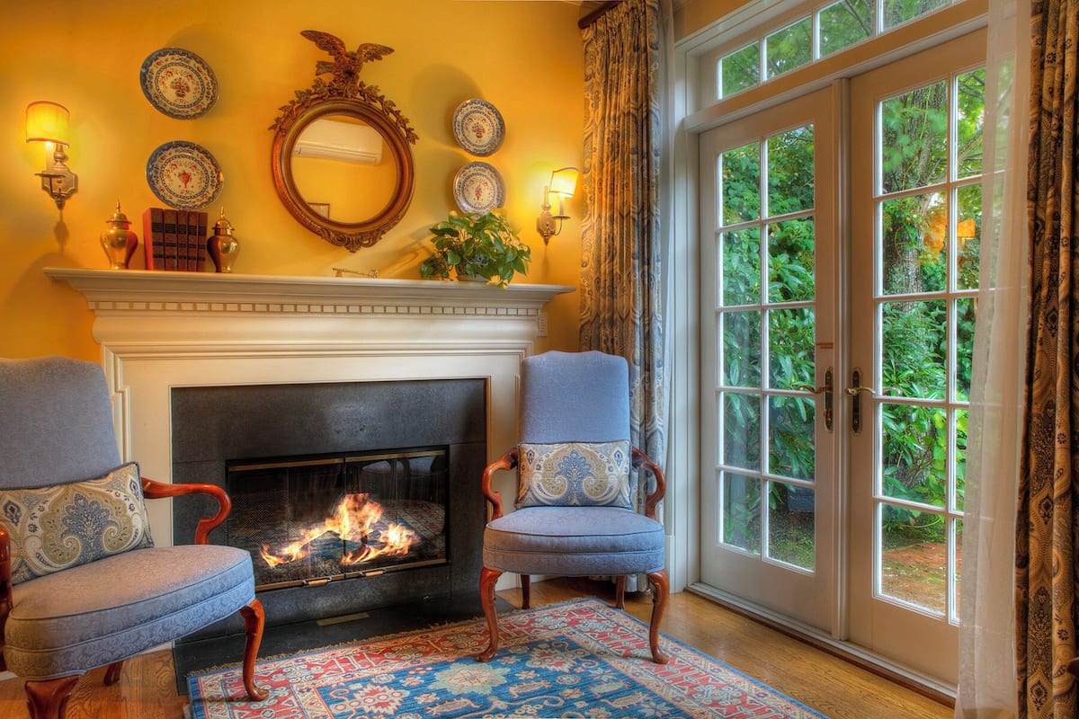 A cozy room with a fireplace, two armchairs, a patterned rug, and a large window with a view of greenery.