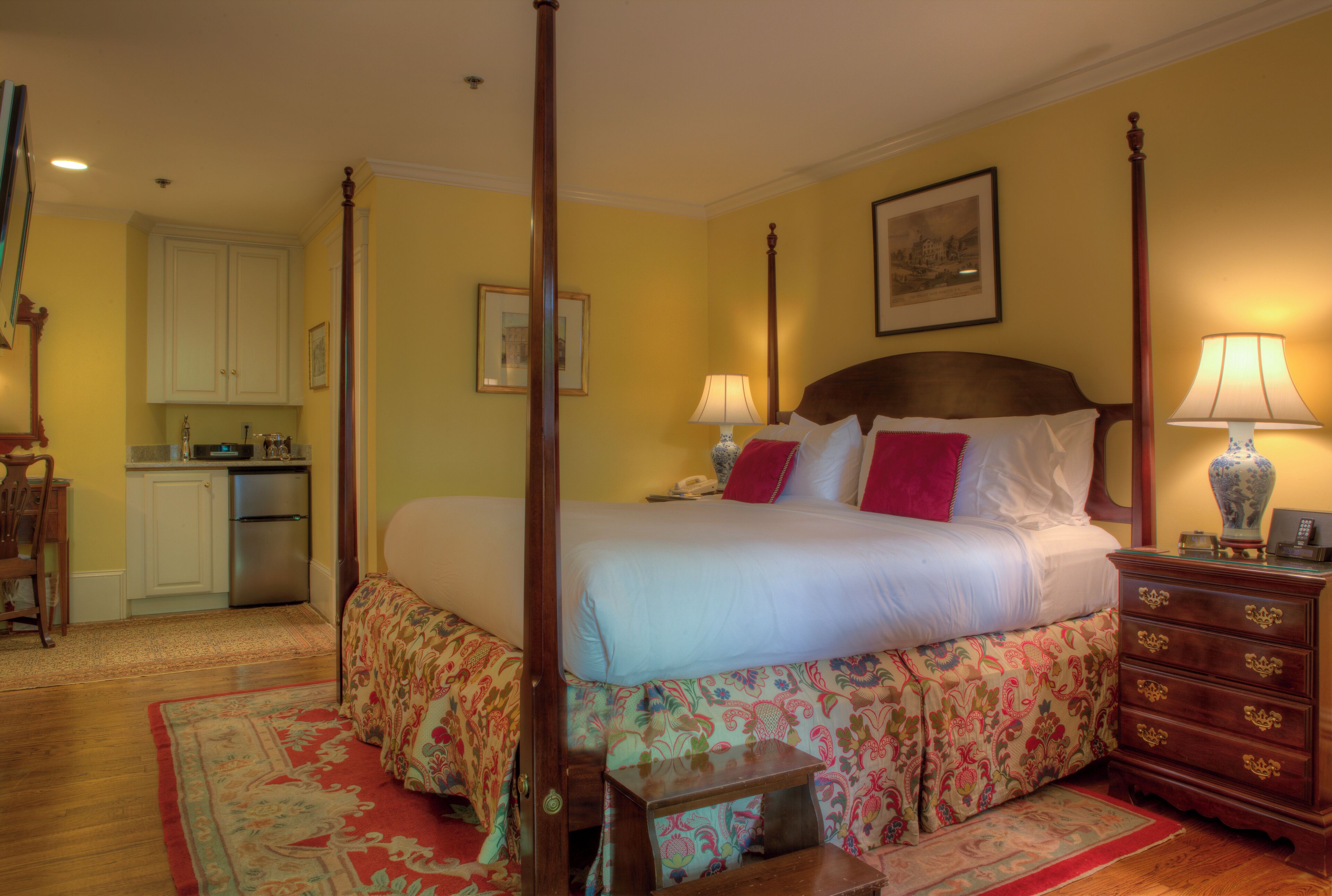 A four-poster bed with a floral bedspread, a wooden nightstand with a lamp, and a small table with a chair.