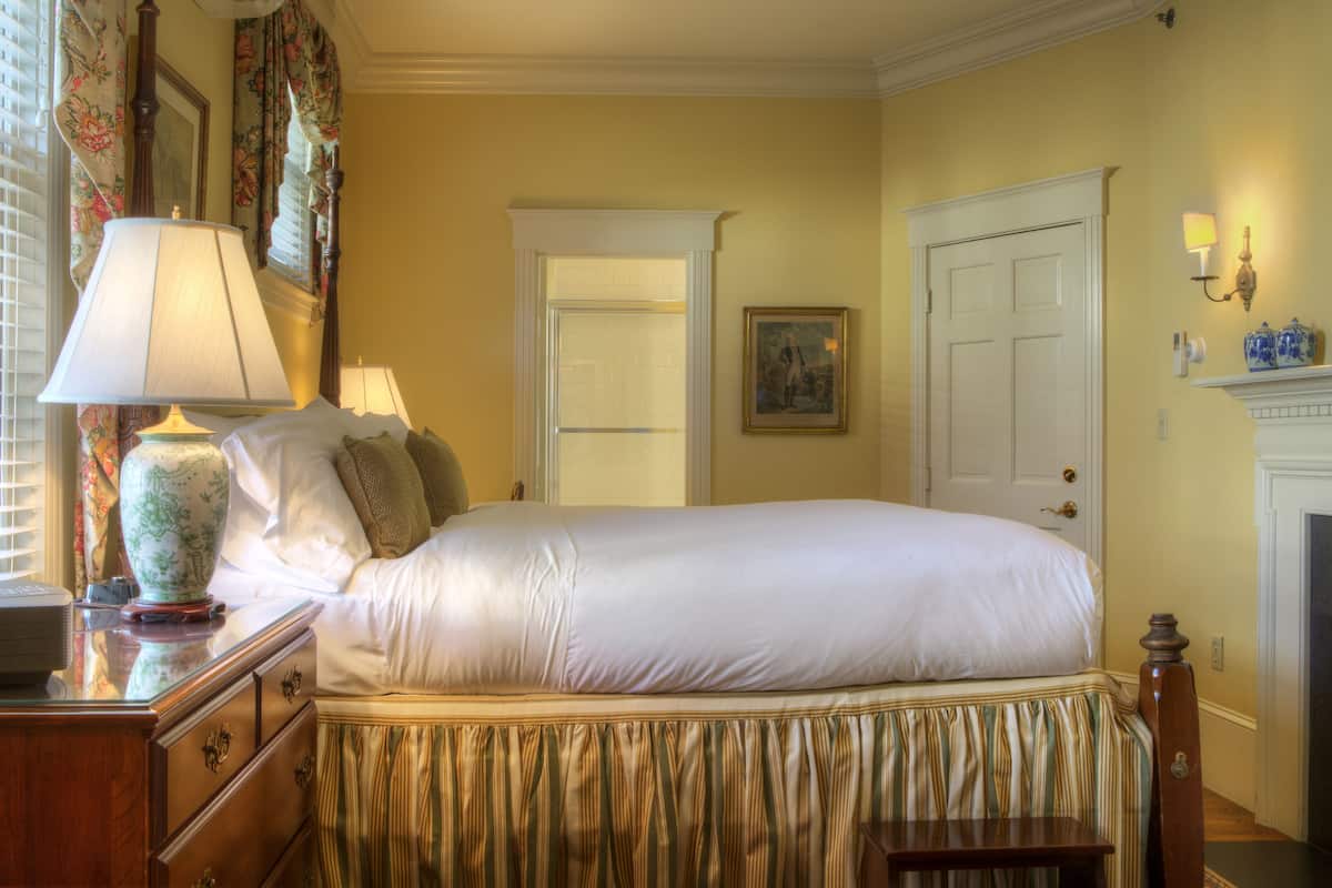 A neatly made bed with a white comforter and a striped skirt, a wooden dresser with drawers, a vase, and a lamp on the dresser.