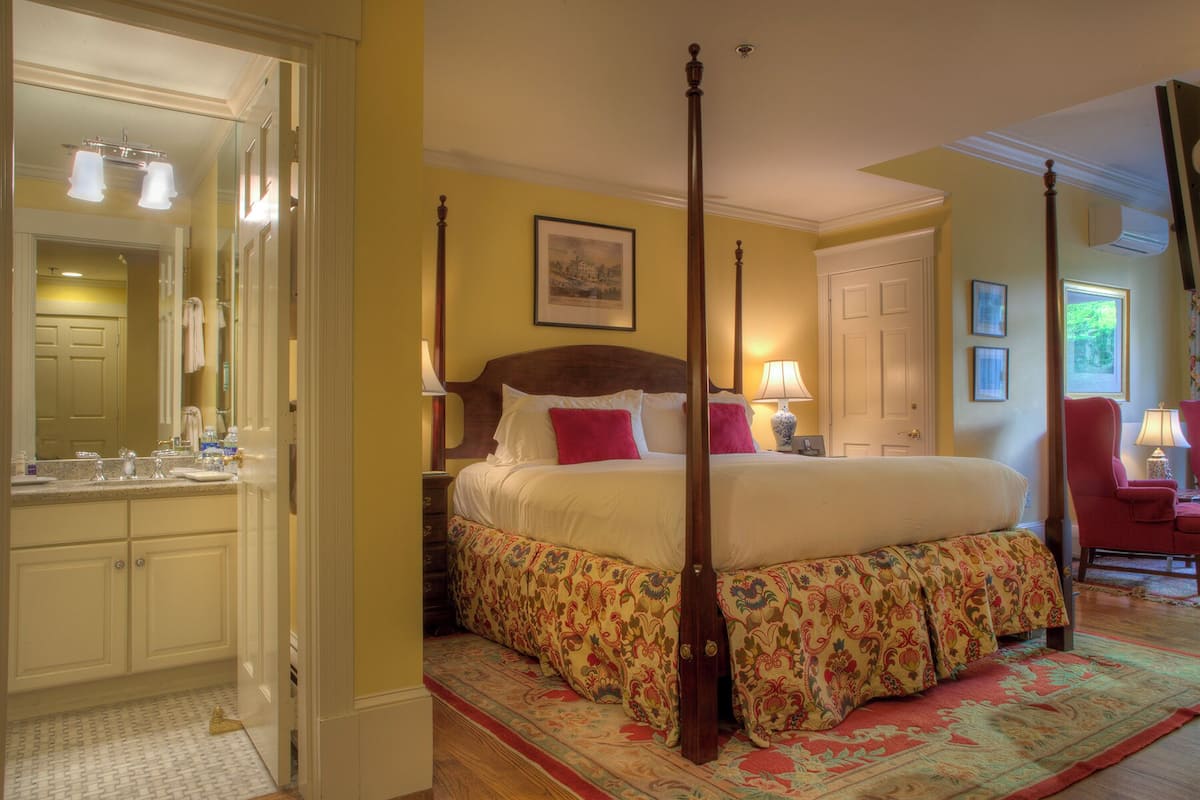 A four-poster bed with a floral bedspread, a bedside table with a lamp, and a large mirror in a room with yellow walls and a wooden floor.