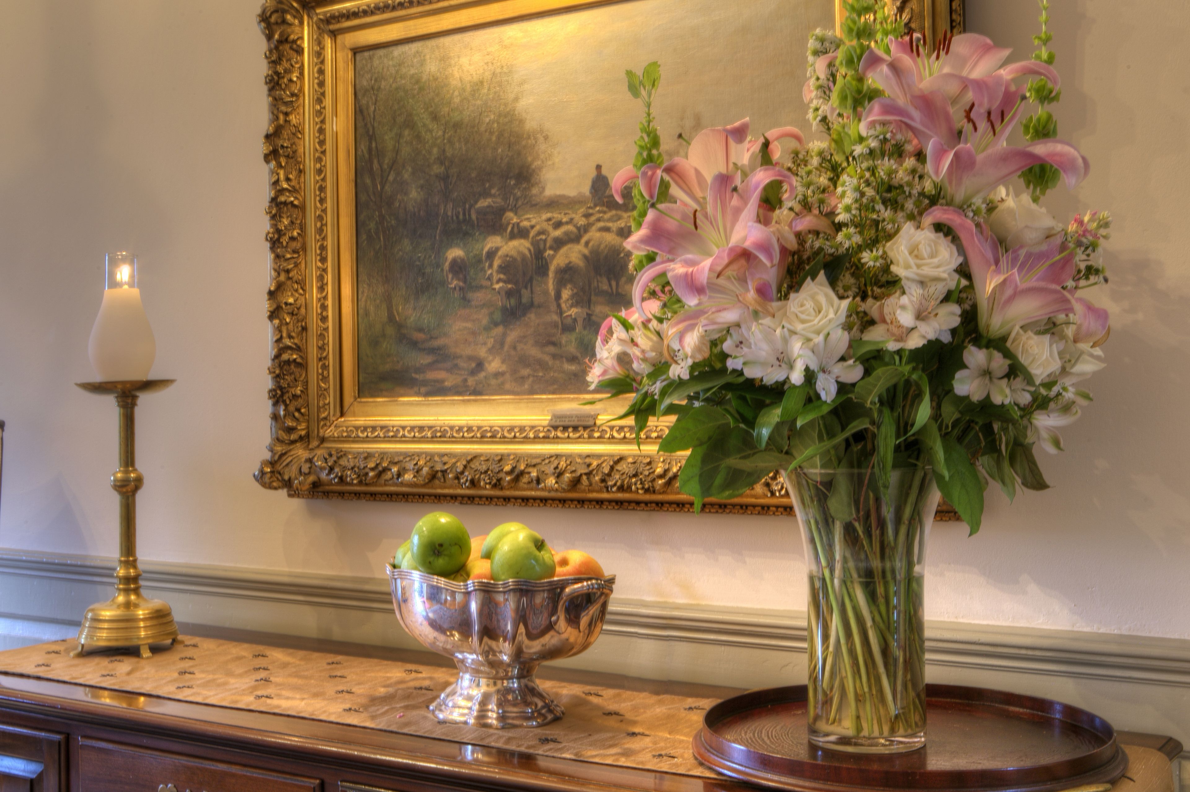 Une table sur laquelle se trouvent un saladier en argent rempli de fruits, un vase de fleurs et une bougie, le tout posé sur une surface en bois.