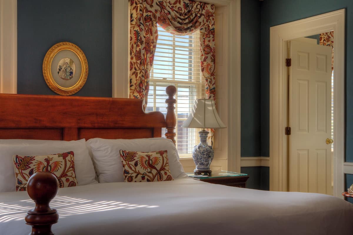 A bedroom with a wooden headboard, a bed with white linens, floral-patterned pillows, a vase on a nightstand, and a door with a brass doorknob.