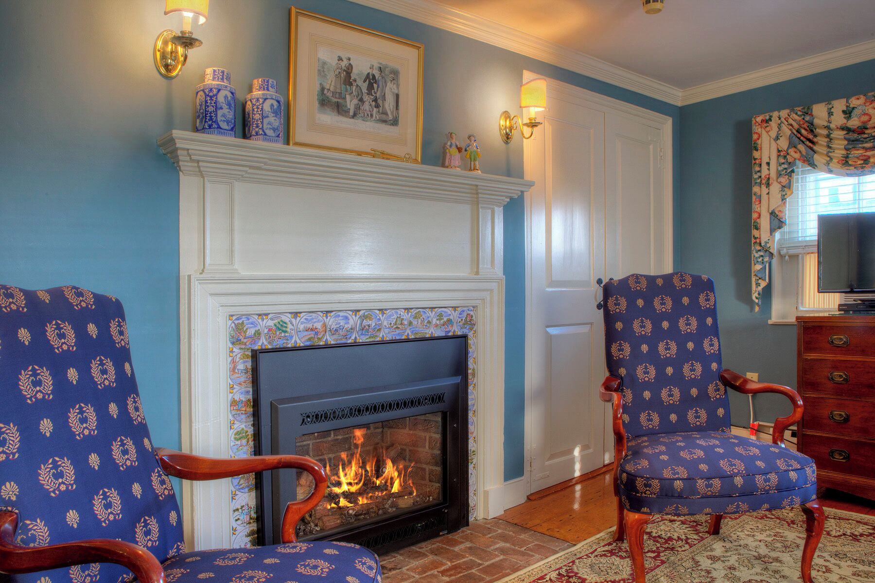 A living room with a fireplace, two patterned chairs, a wooden dresser, and a television.