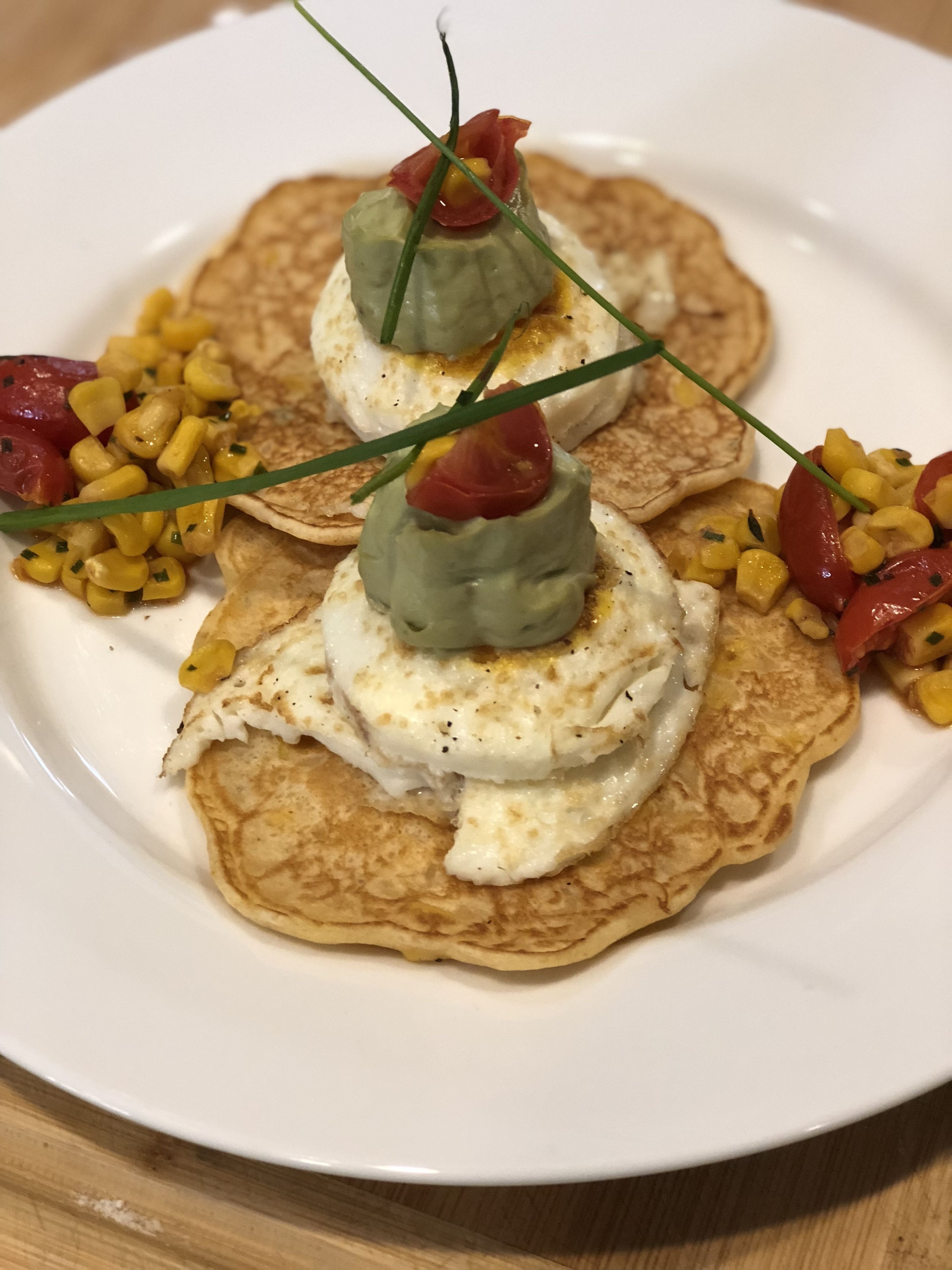 Une assiette de crêpes garnies d’une cuillerée de crème et d’une garniture verte, accompagnée d’une portion de maïs et de tranches de tomates.