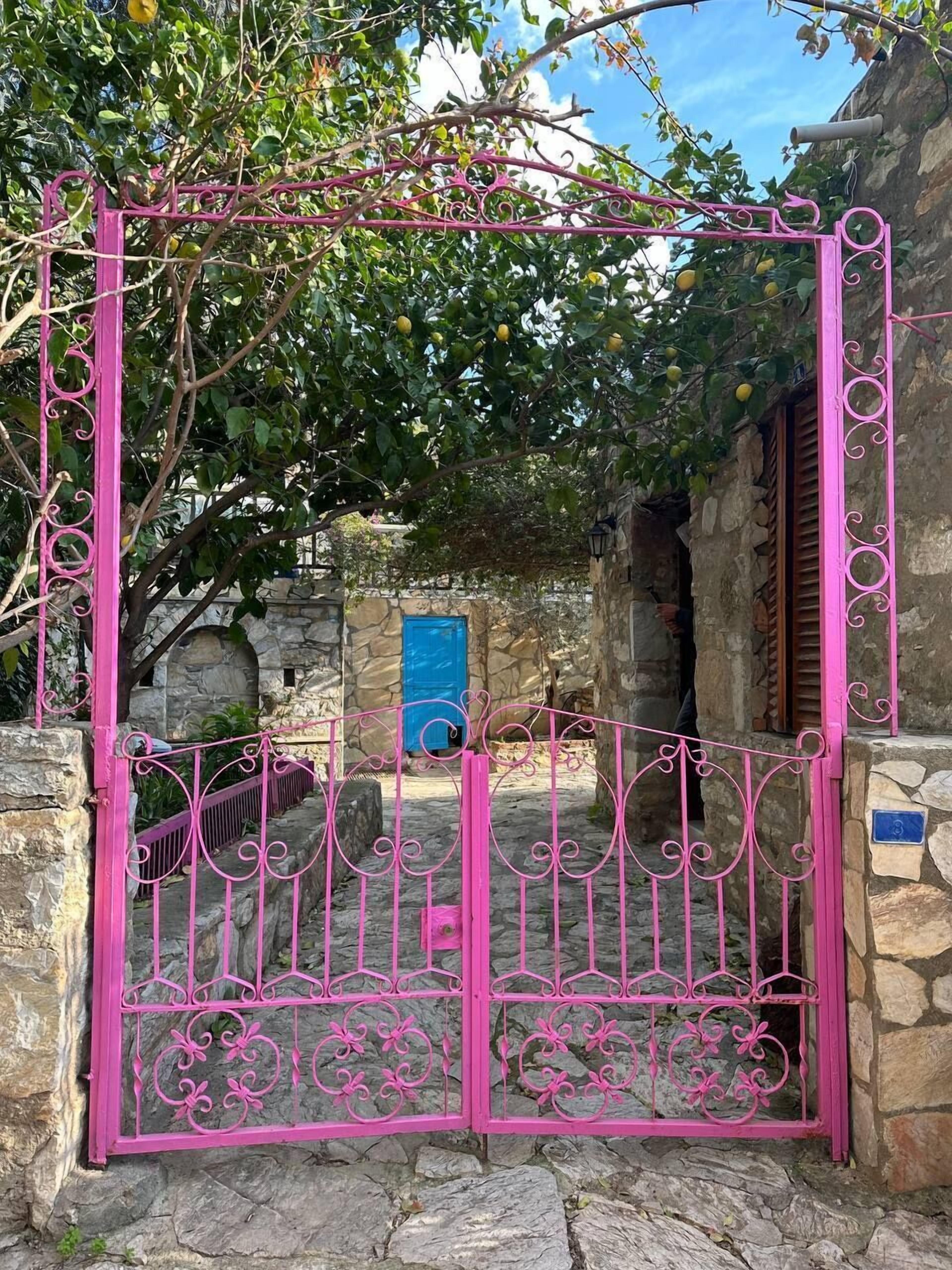 Een roze metalen poort met decoratieve patronen, die leidt naar een stenen gebouw met een blauwe deur.