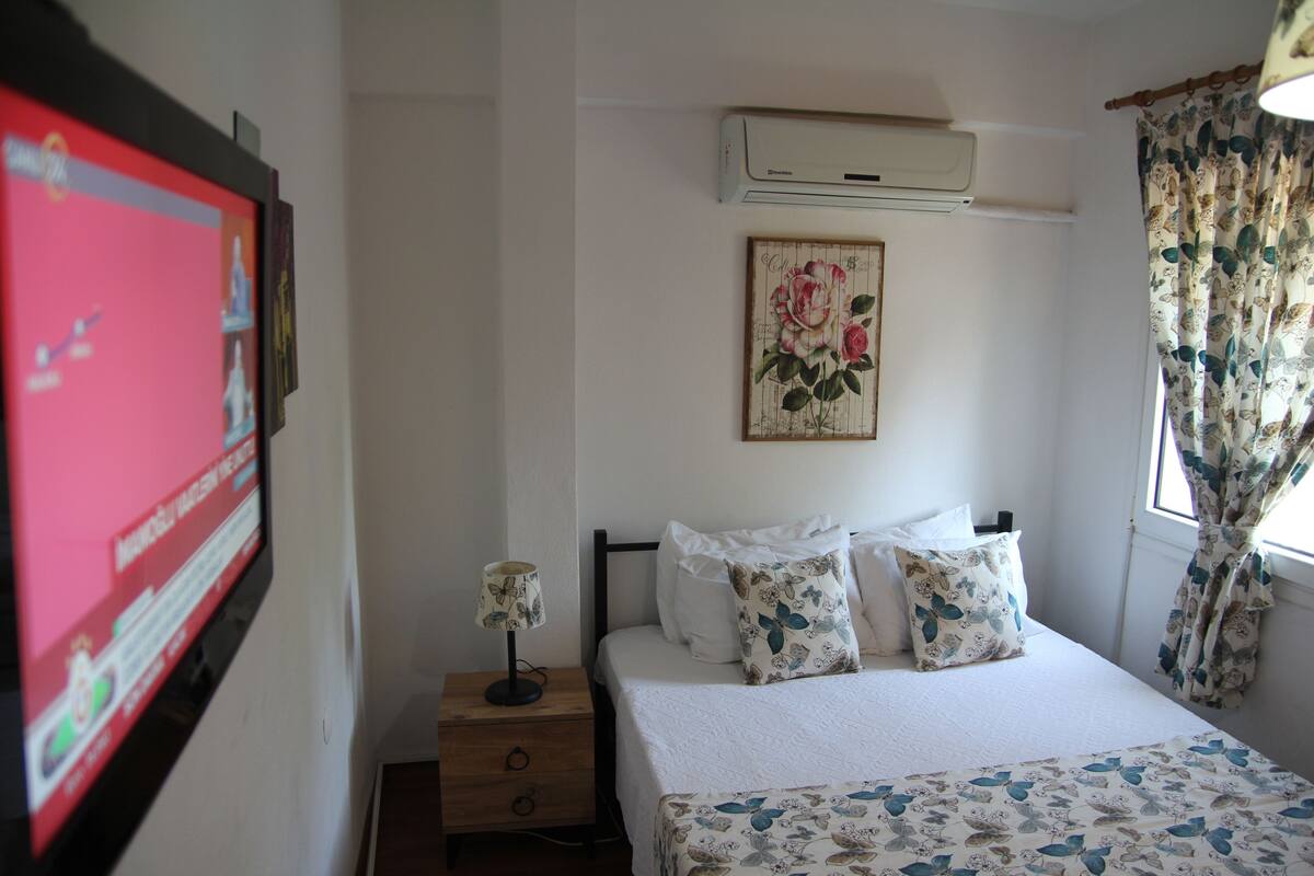 A bedroom with a bed, a nightstand, a wall-mounted air conditioner, a framed picture, and a window with curtains.