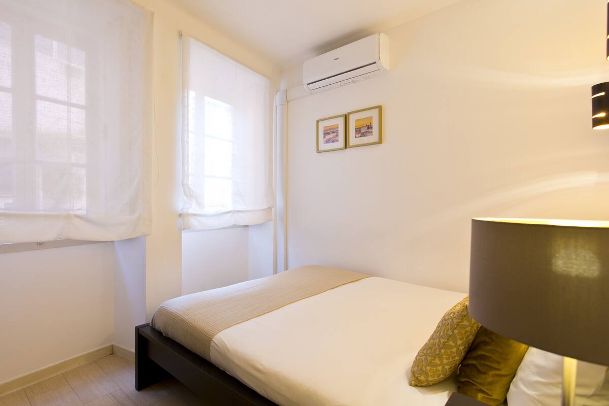 Een slaapkamer met een bed, een raam met gordijnen, een airconditioning en een wandlamp.