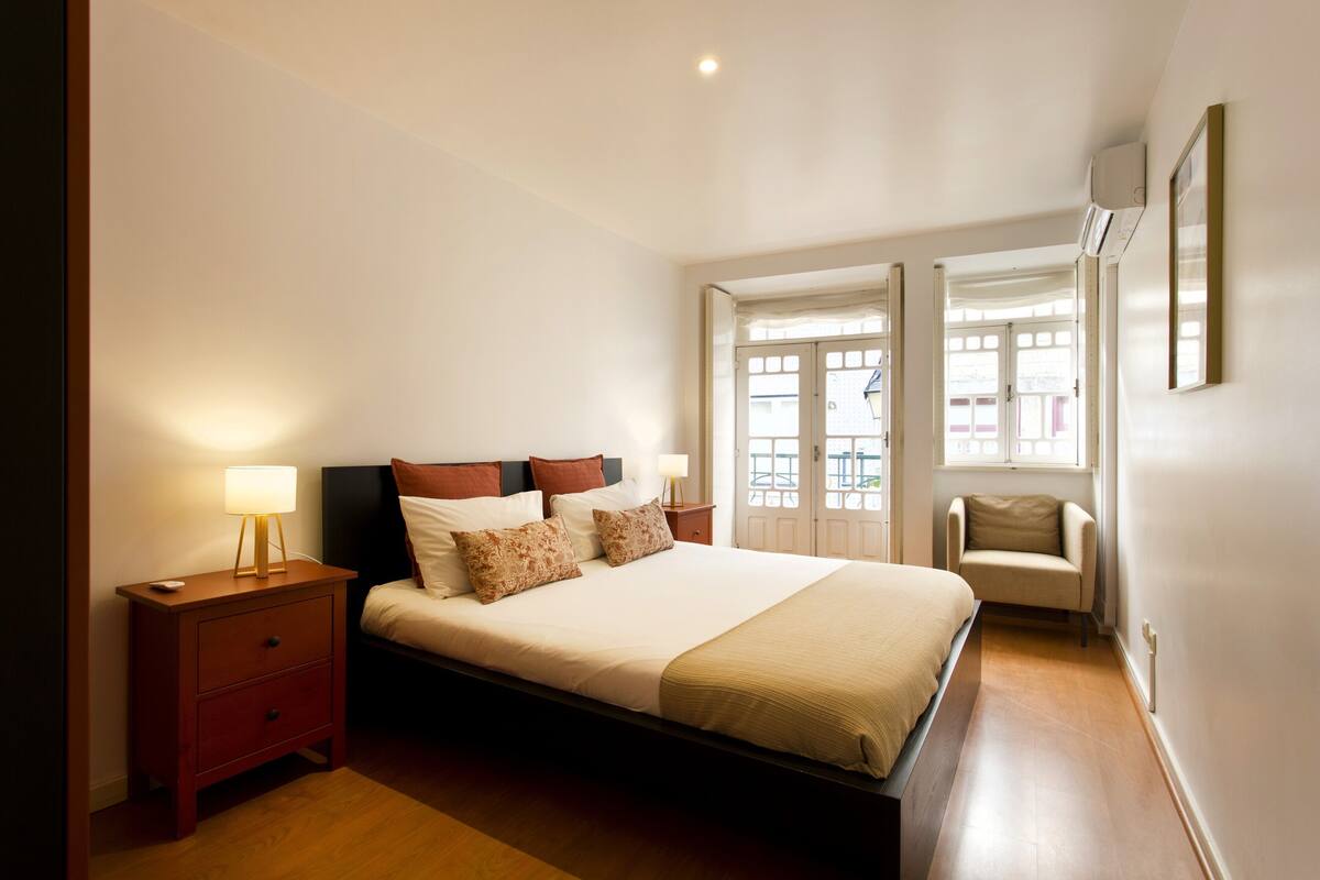 Een slaapkamer met een bed, nachtkastjes, een lamp, een stoel en een raam met uitzicht.